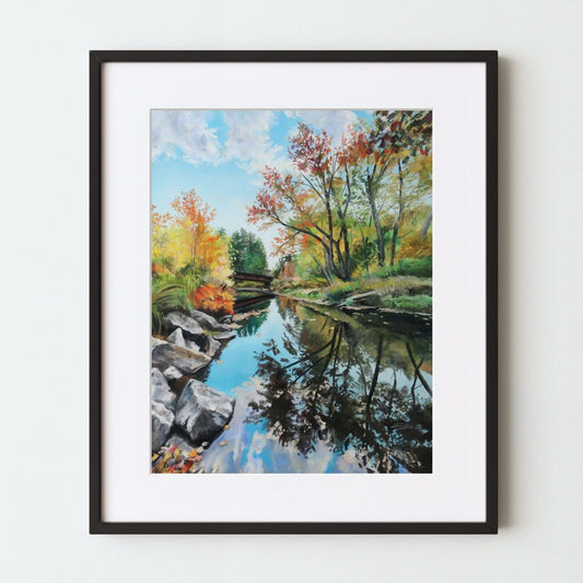 A mockup of a landscape art print in a black frame with a white mat. The painting shows a still river at the bottom with a bridge further away, surrounded by fall foliage and a blue sky. The reflection in the water is compelling.