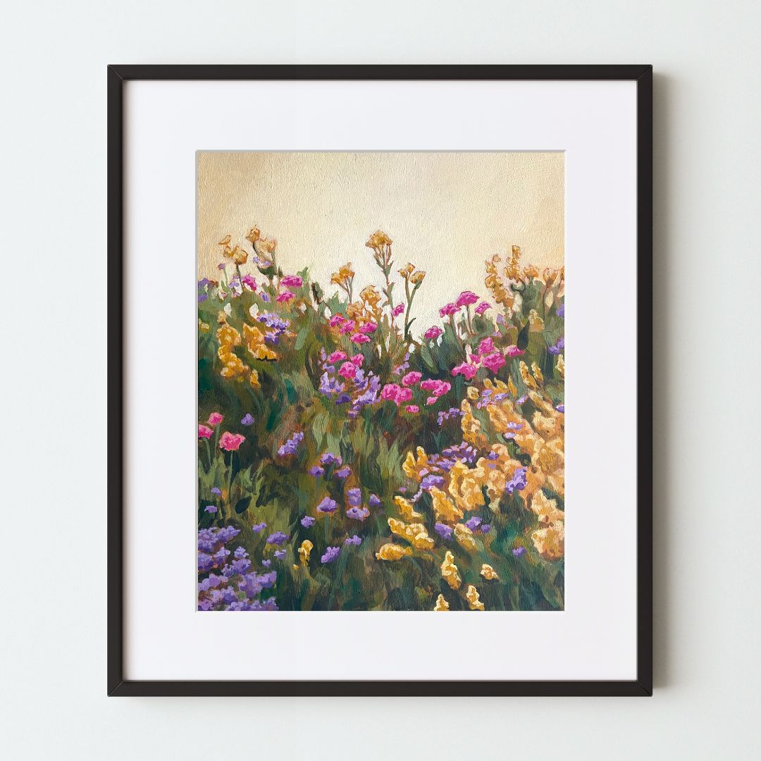 A mockup of a floral art print in a black frame with a white mat. The painting shows lush green grass with pink, purple, and golden flowers in gentle movement together. They all reach up toward a soft yellow sky.