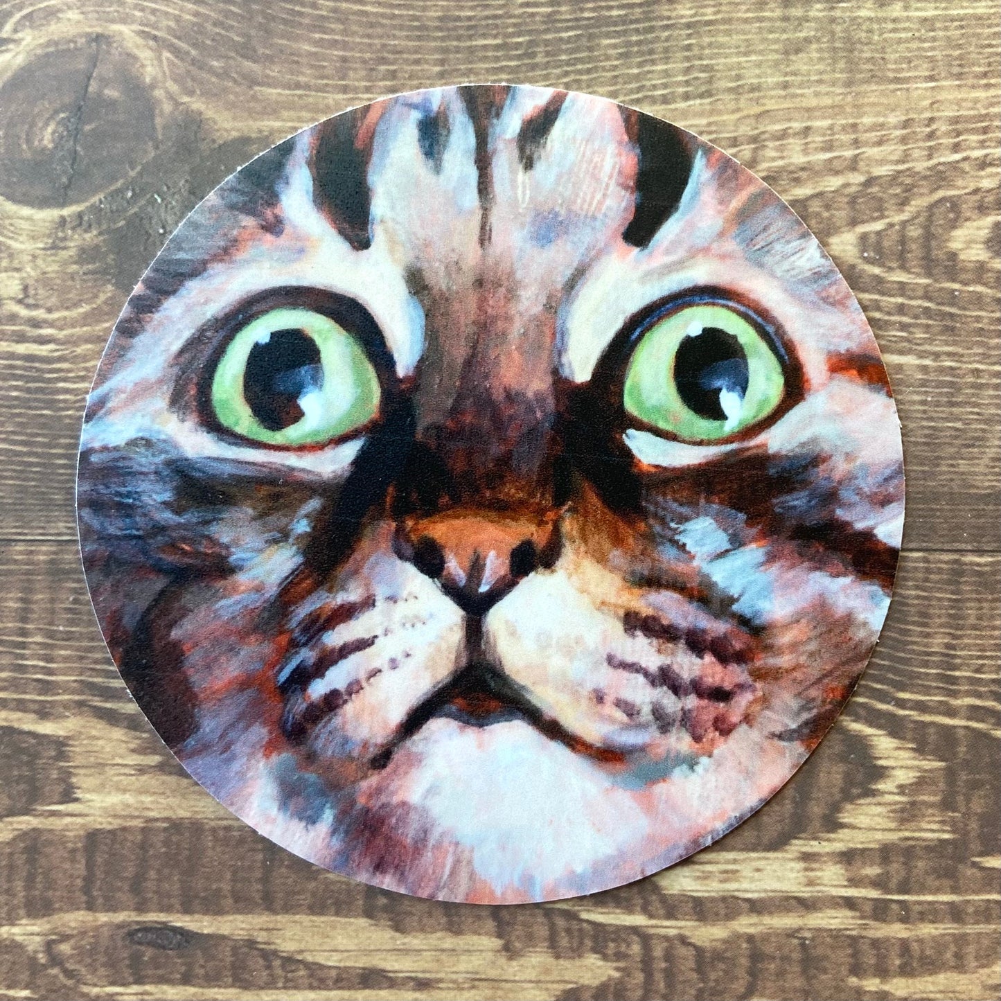 A round sticker of my painting "Alert Eyes", showing a gray tabby cat with bright green eyes, close-up so the face fills the frame. The sticker is sitting on a dark wood surface. The painterly style of the feline art shows in this decal reproduction.