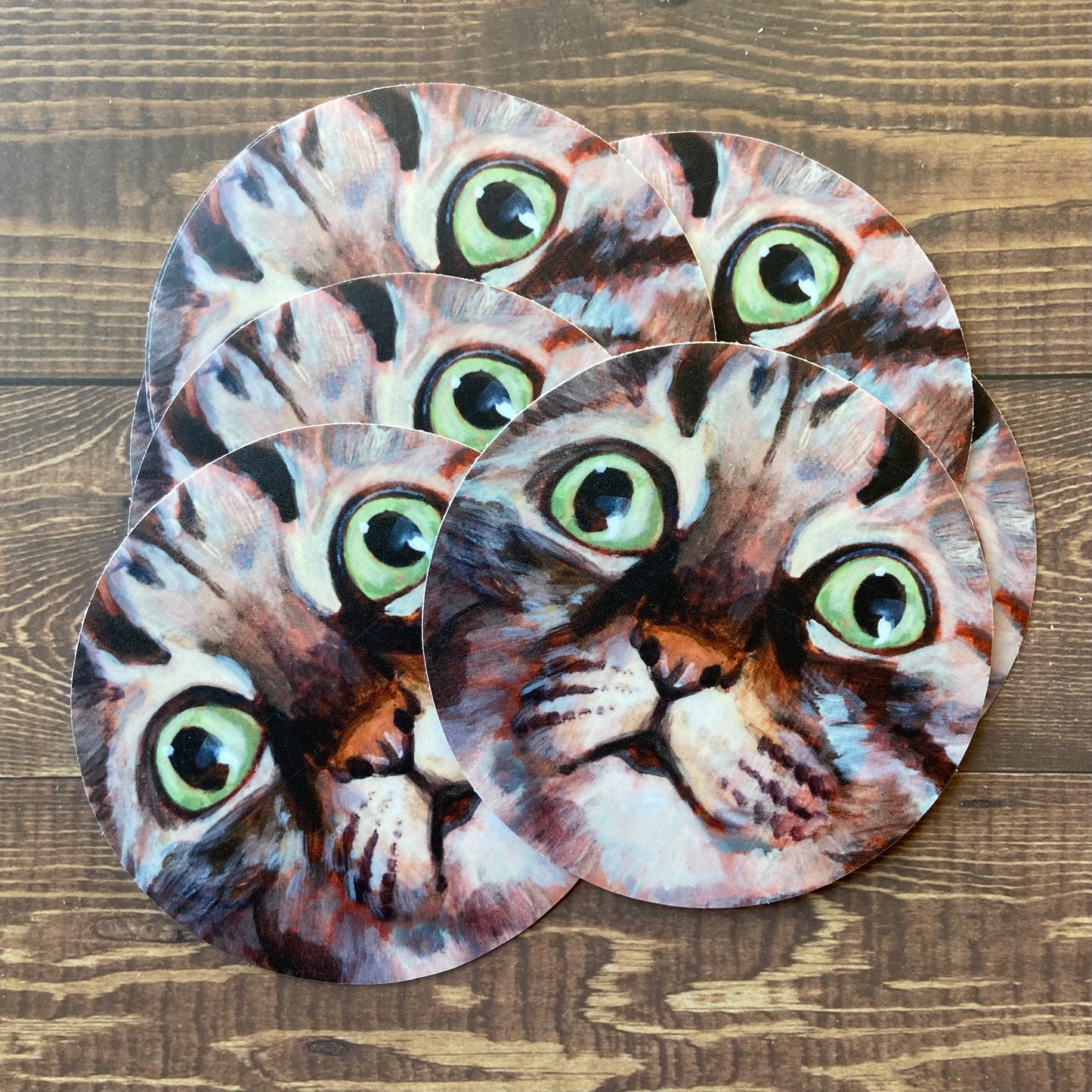 A small pile of round stickers of my painting "Alert Eyes", showing a gray tabby cat with bright green eyes, close-up so the face fills the frame. The feline's expression looks as though she's hunting, and the paint brush strokes look like fur.