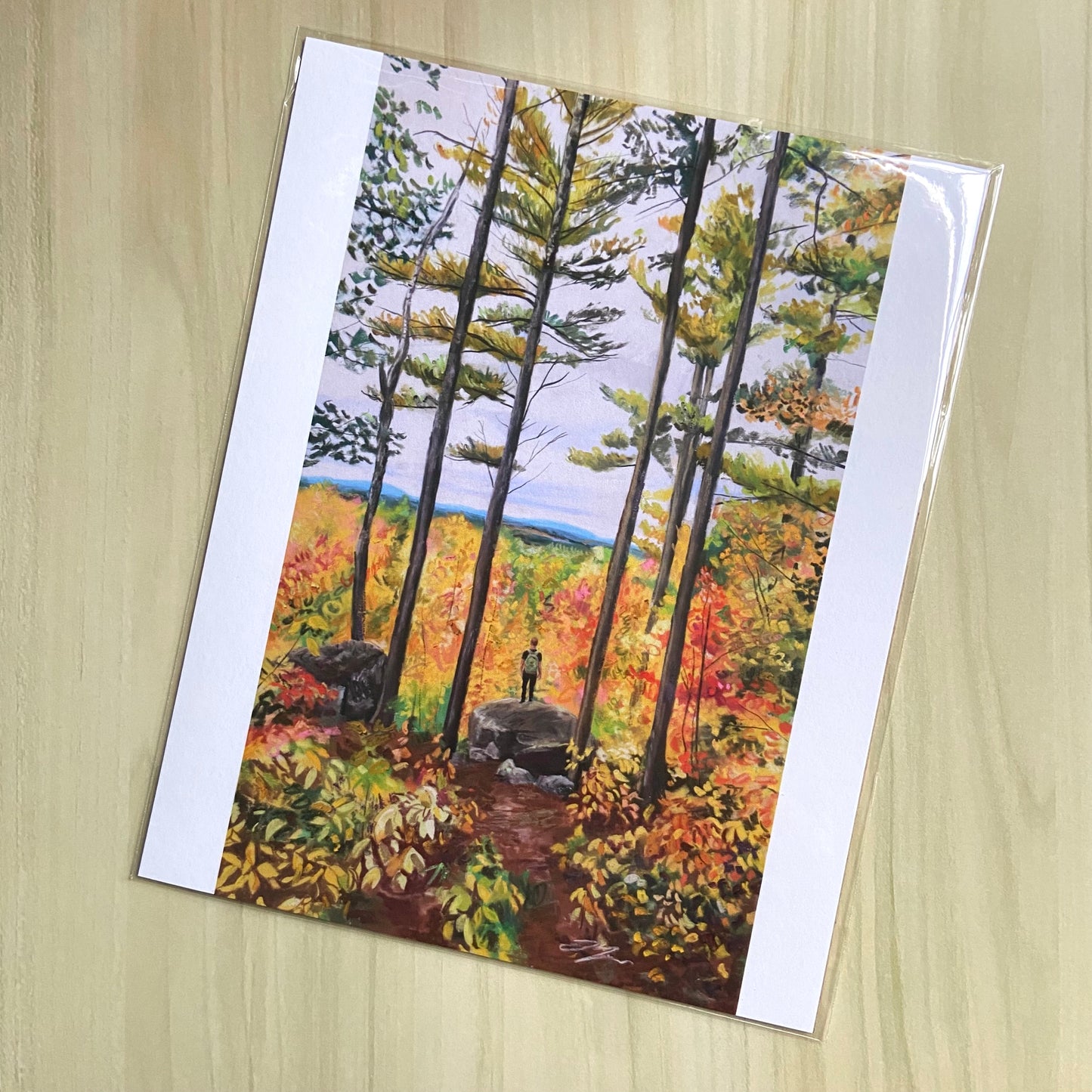 An art print of an autumn forest scene. A man stands on a rock in the woods, looking out at fall foliage and distant mountains. He is surrounded by tall pines and low brush in fall colors. The print has white borders on the side edges. 