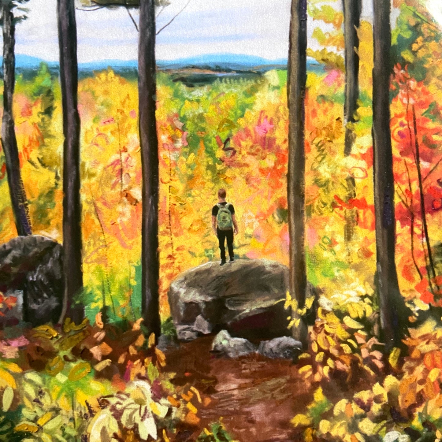 A close-up detail of the wall art print. The hiker is shown here, a man dressed in black with a green backpack. He stands on a boulder in the forest with fall colors all around. Past yellow trees are distant blue mountains, the sky overcast.