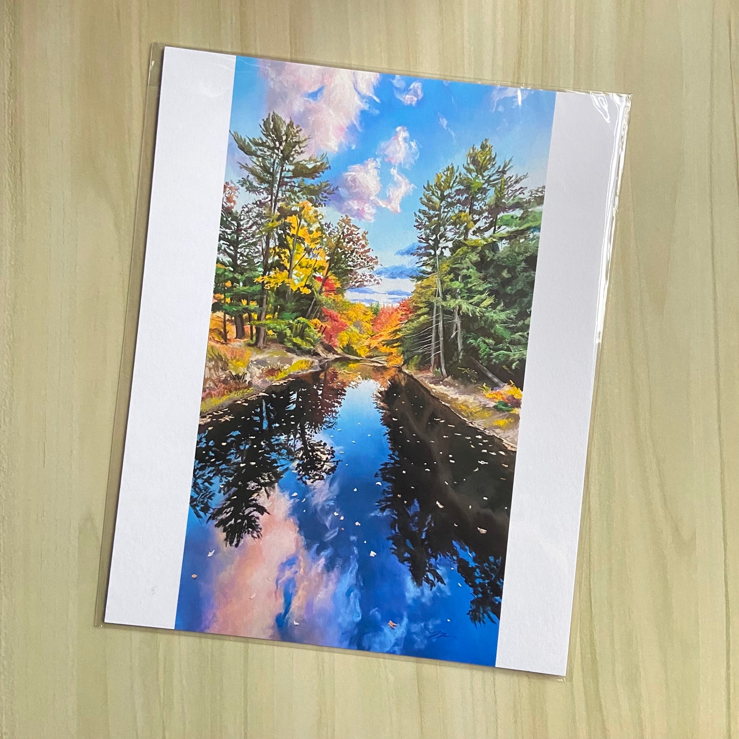 An art print of a river running under the viewer and to the center of the image. On either side are tall trees, some evergreen and some with fall colors. The blue sky and pink clouds are reflected in the water. The print has white side borders.