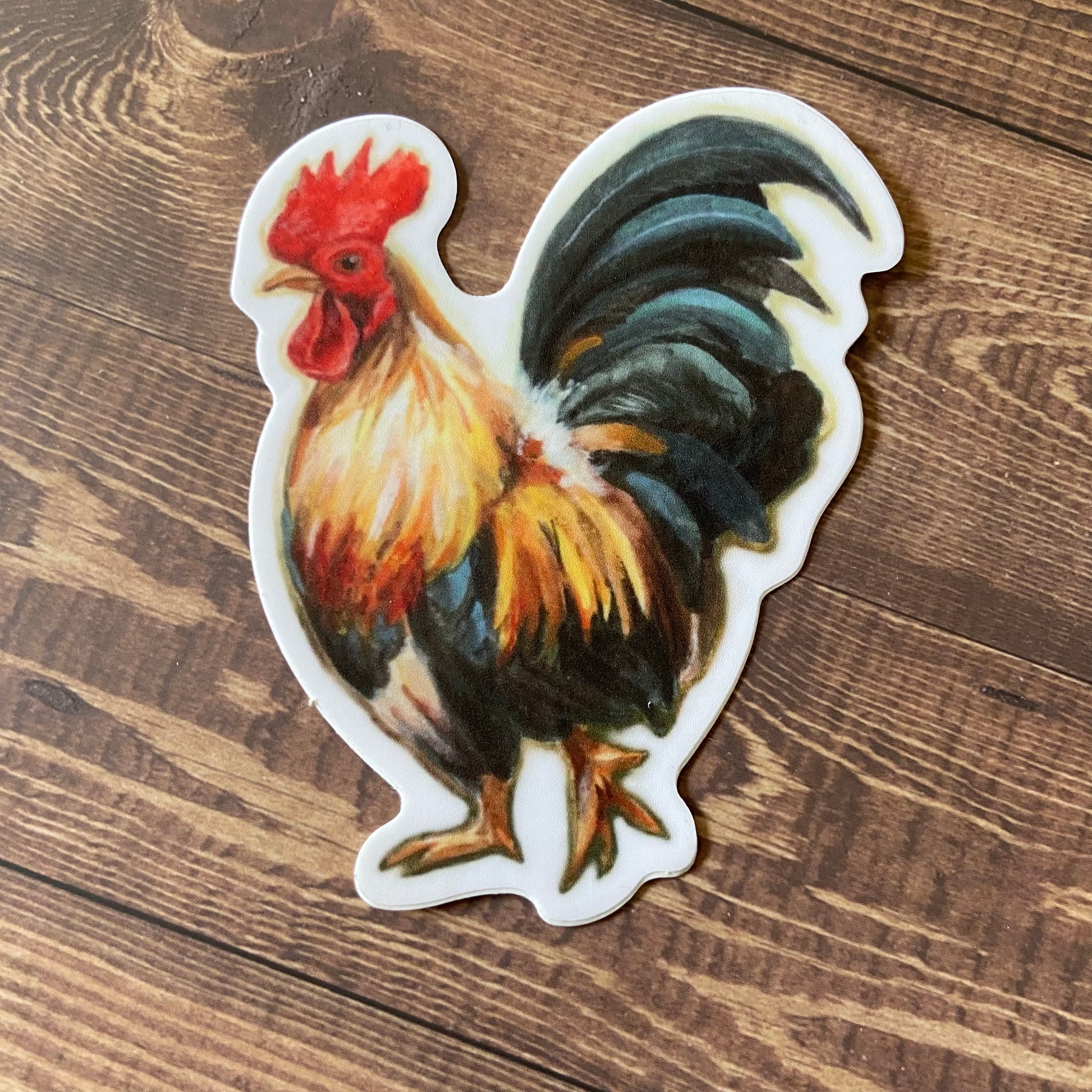 A sticker shaped like a rooster sitting on a wooden surface. The decal is a painting of a bantam rooster in profile, with feathers in orange/yellow and blue/black. He is mid-stride and looking at the viewer. Sticker has a thin white border.