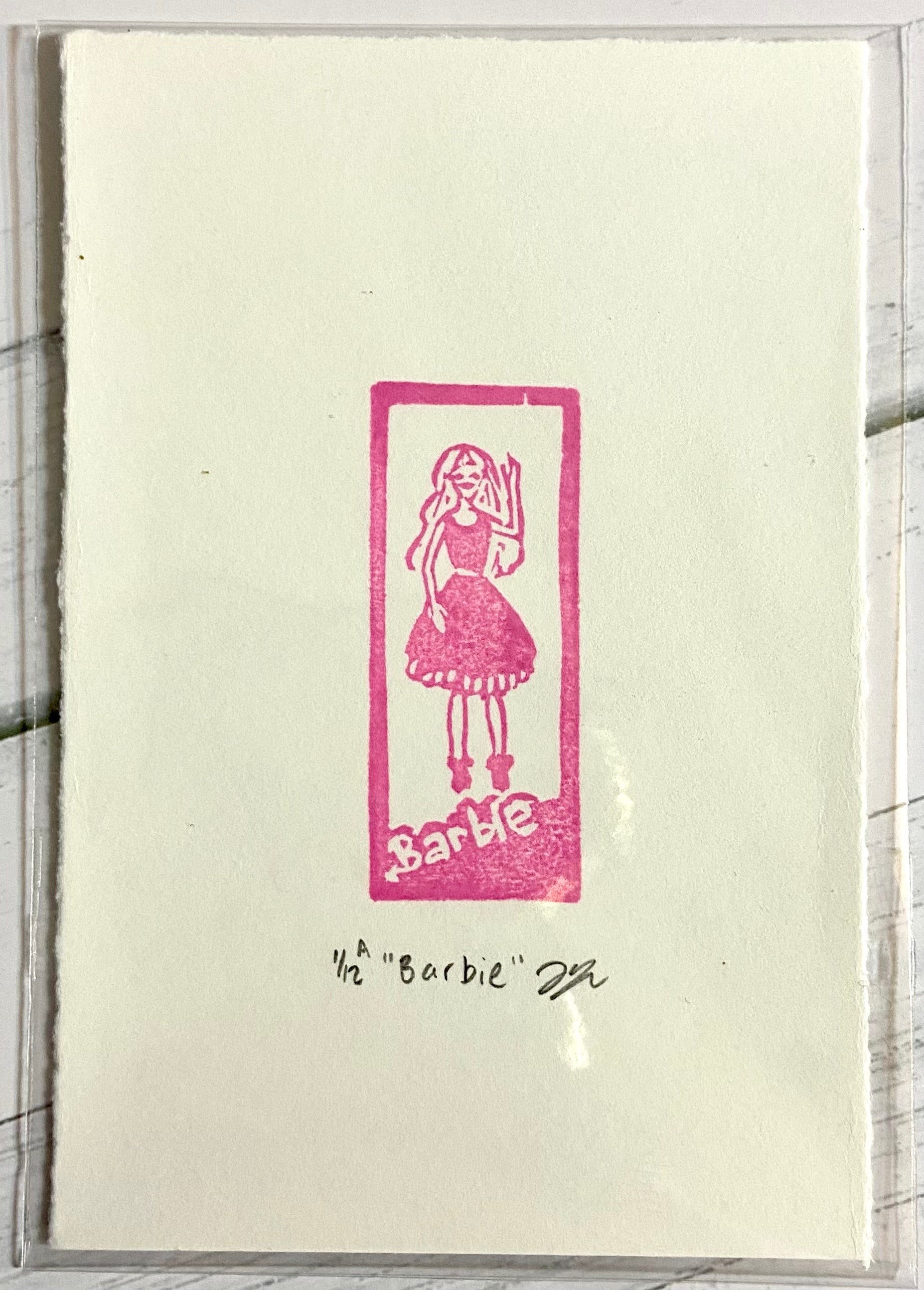 A pink relief print depicting a Barbie in a box. The print is made by hand with a carved pink eraser and is numbered, signed and titled.