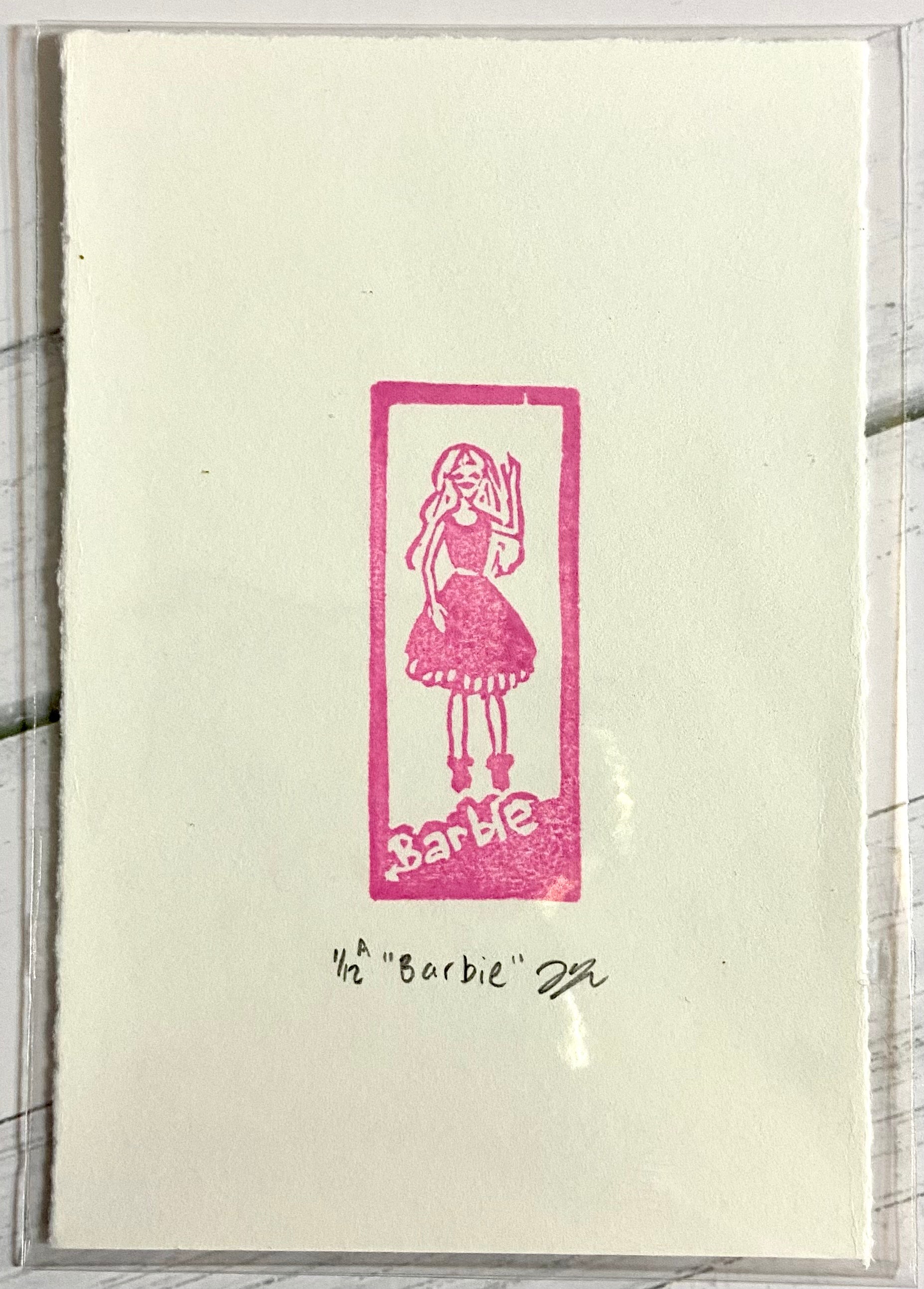 A pink relief print depicting a Barbie in a box. The print is made by hand with a carved pink eraser and is numbered, signed and titled. 