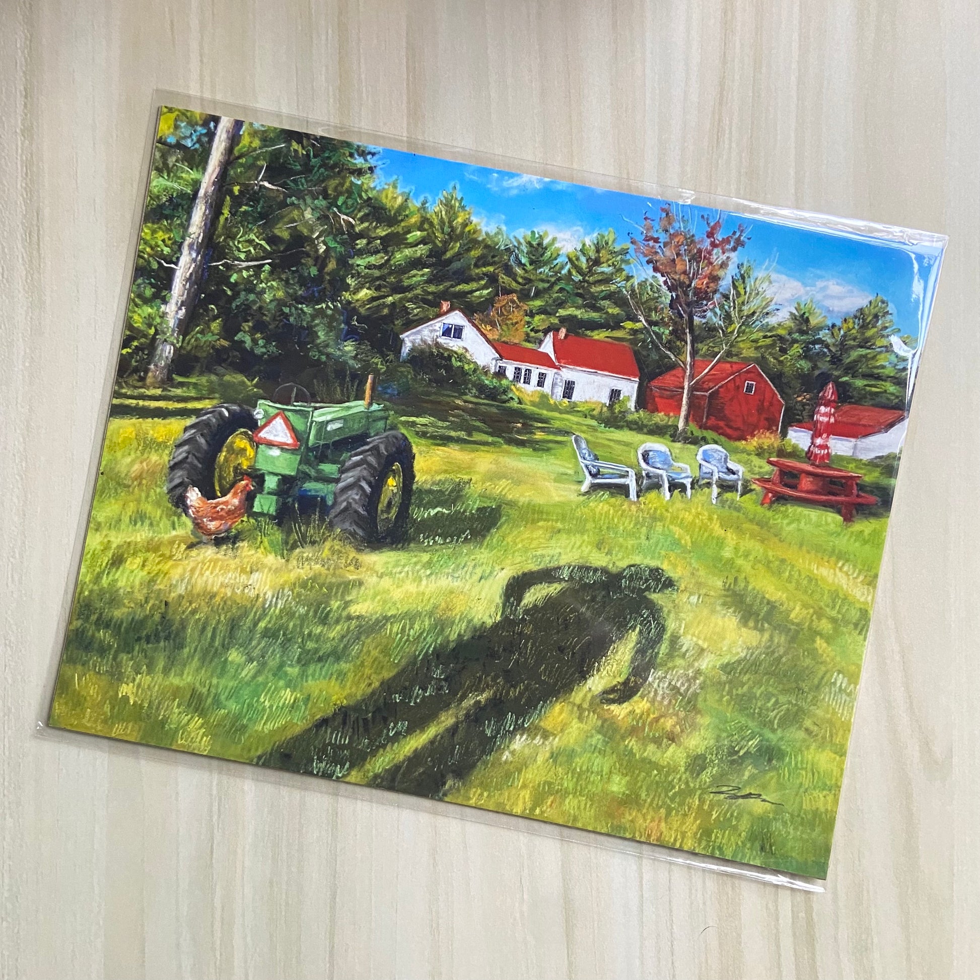 An art print of a farmhouse scene painting. A green grass field is most of the painting, with a long shadow of a man in front, and a green tractor, chicken, and picnic table nearby. In the distance is a white farmhouse, red barn, and green forest.