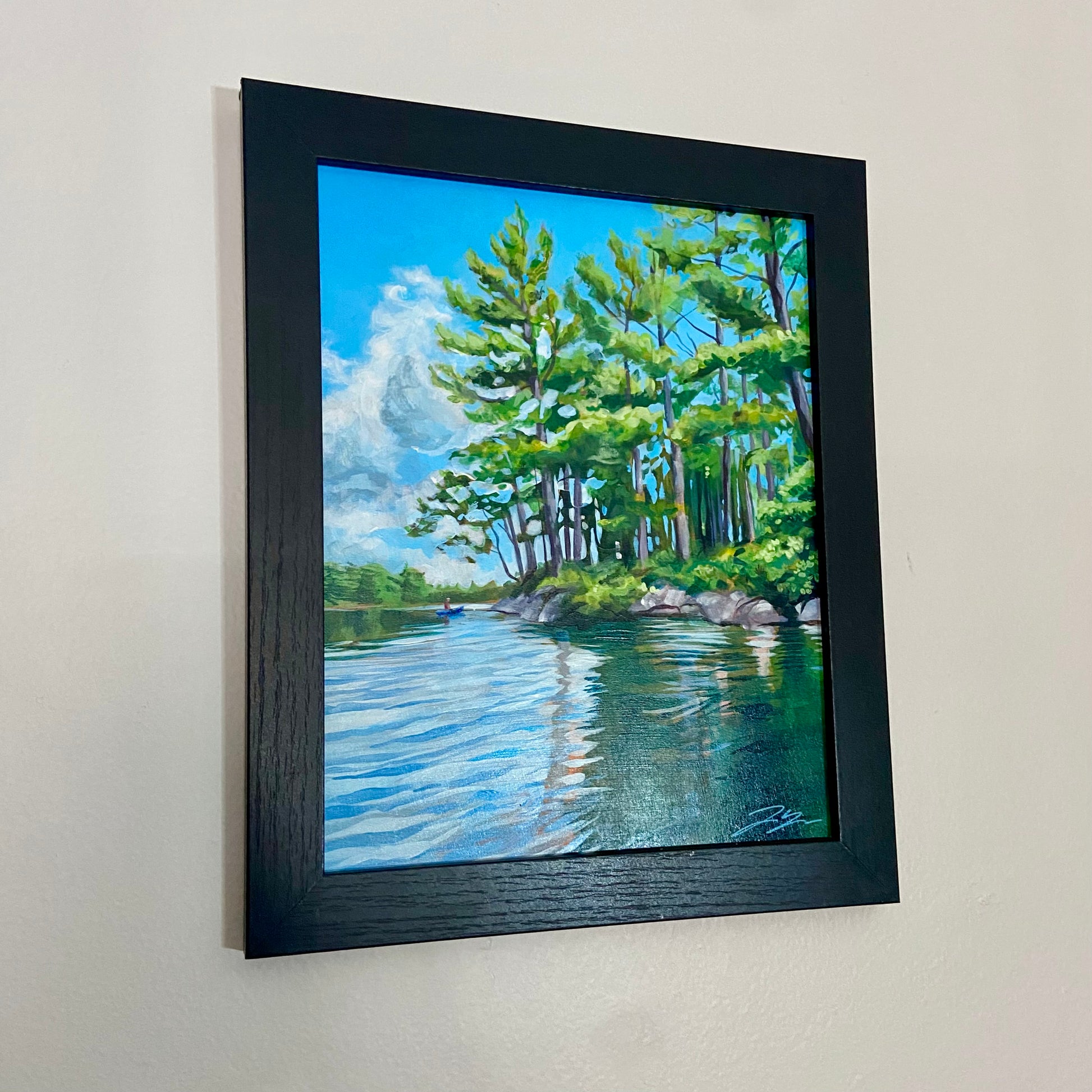 A painting, mainly light blue and green, of some trees at the edge of land, viewed from the surface of a lake. There is a kayaker far away near the land. The painting is in a black wood frame and hangs on a light gray wall. 