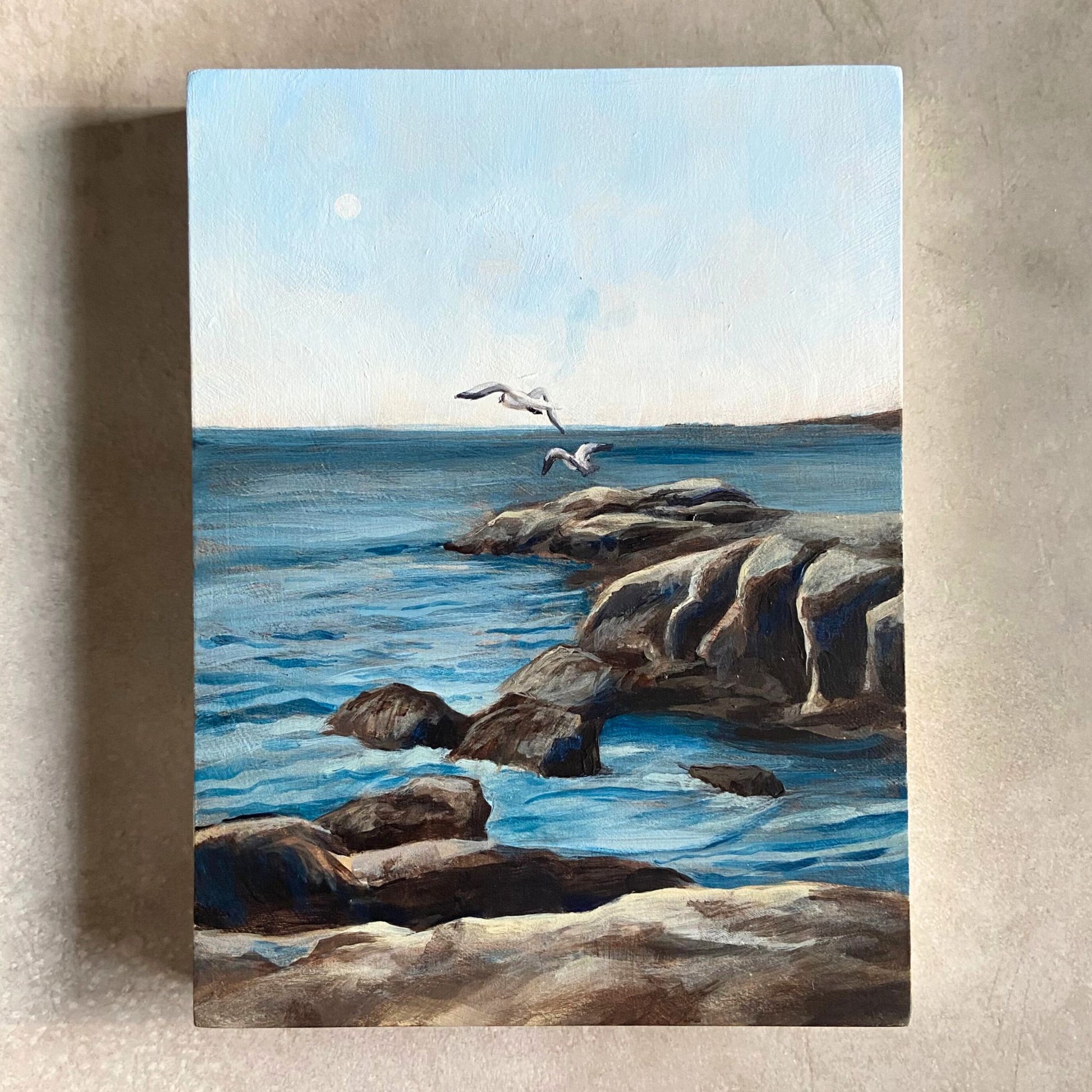 A seascape painting on wood panel sitting on a beige, textured surface. The painting, portrait oriented, depicts a rocky area of blue water with the horizon in the distance. Two seabirds are flying around the center of the piece, at horizon level. 
