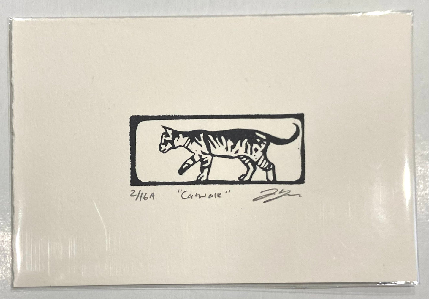 A black and white relief print of a striped cat walking from right to left, visually enclosed by a box. The prints are made by hand with a carved eraser and numbered, titled and signed.