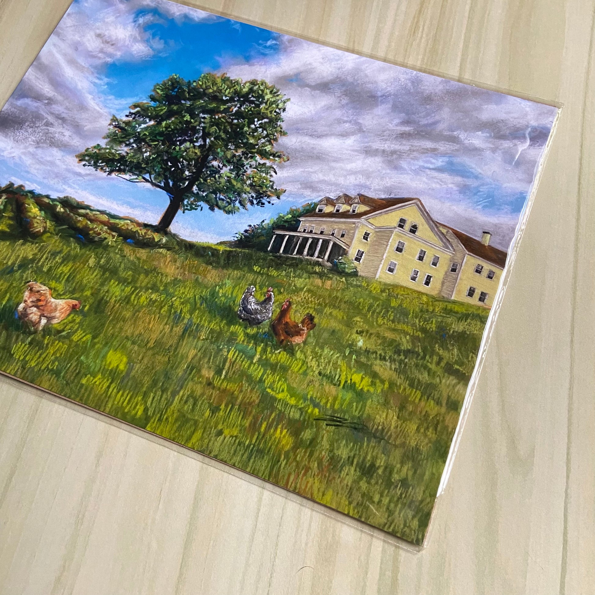 A detail view of the New England/ Maine countryside art print. The soft pastels used to paint the picture are vibrant and have a visible chalky texture. The print shows a yellow home on a green hill under a wide blue sky, with a tree and chickens.