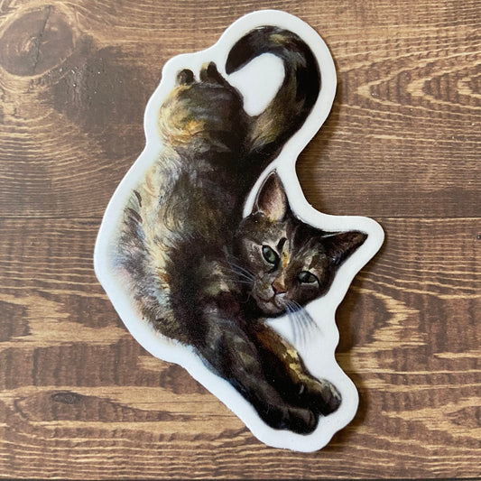 A sticker of a tabby cat laying out, stretching long toward the viewer. The cat's tail is curled upward. The image is painted in watercolor and has a thin white border. The decal is on a dark wooden surface.