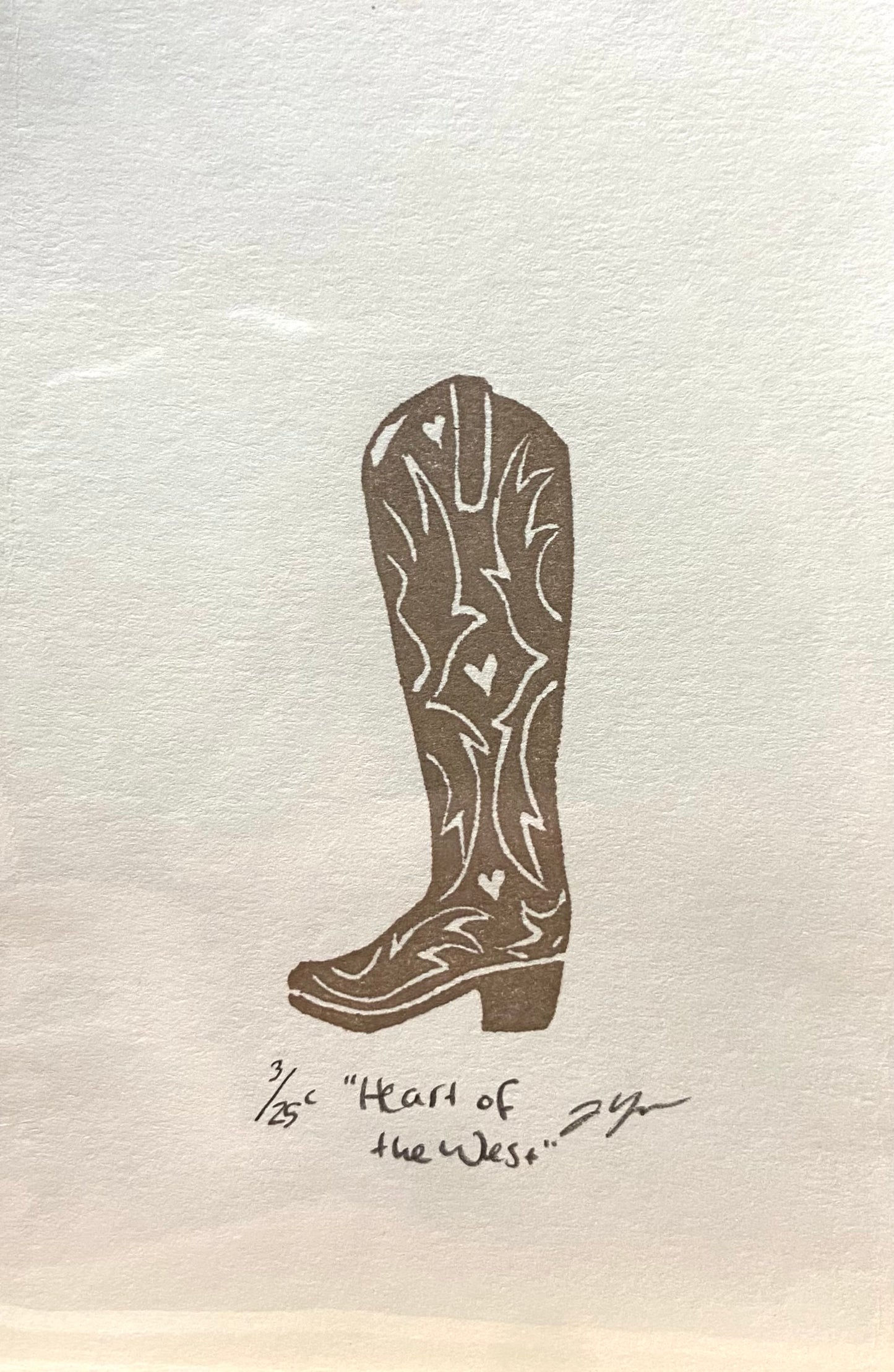 A brown and white relief print of a cowgirl boot, made by hand using a carved eraser. The print is numbered, signed, and titled "Heart of the West".