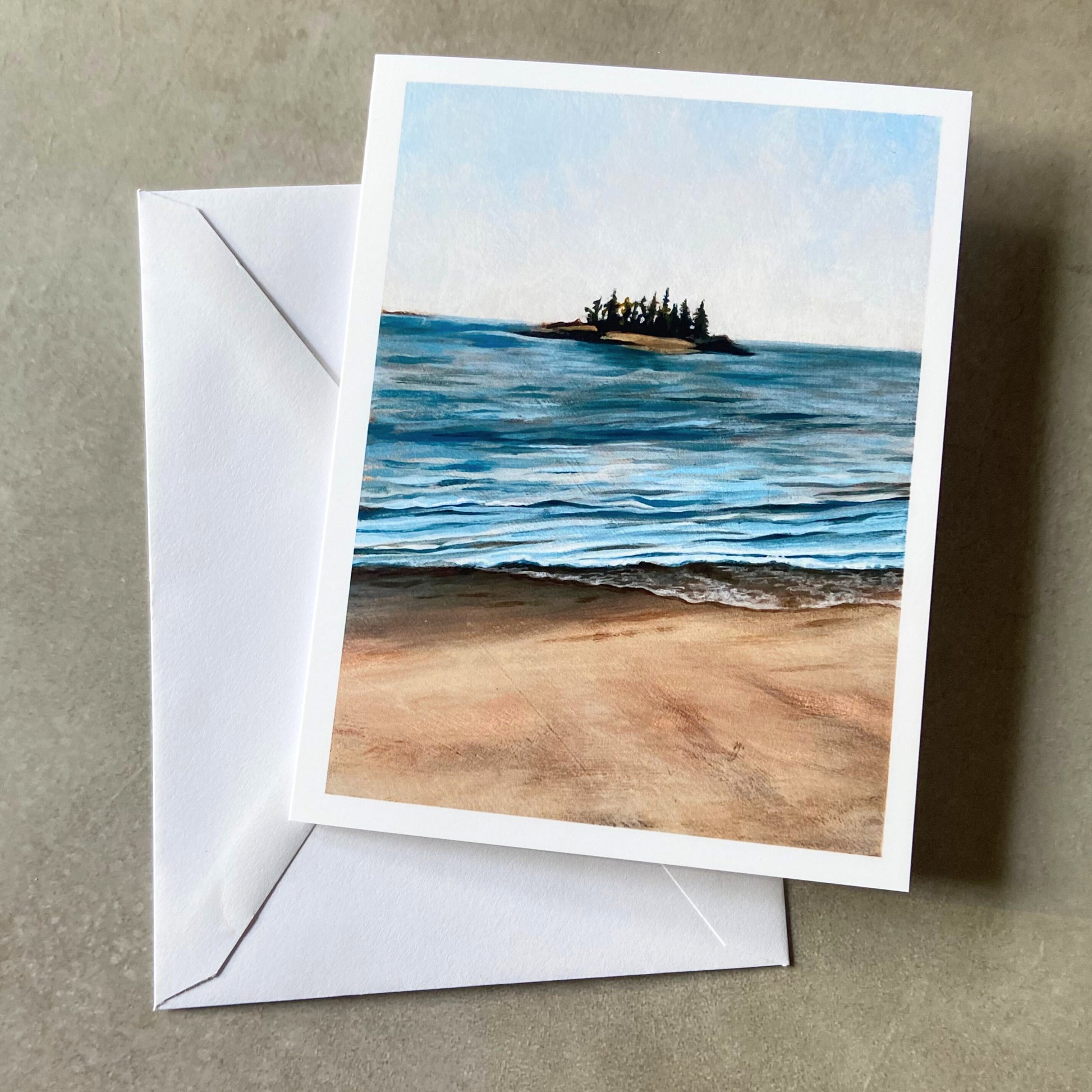 A folded white greeting card and envelope on a grey surface. On the front of the card is a painting showing an island on the ocean horizon and a sandy beach at the bottom. 