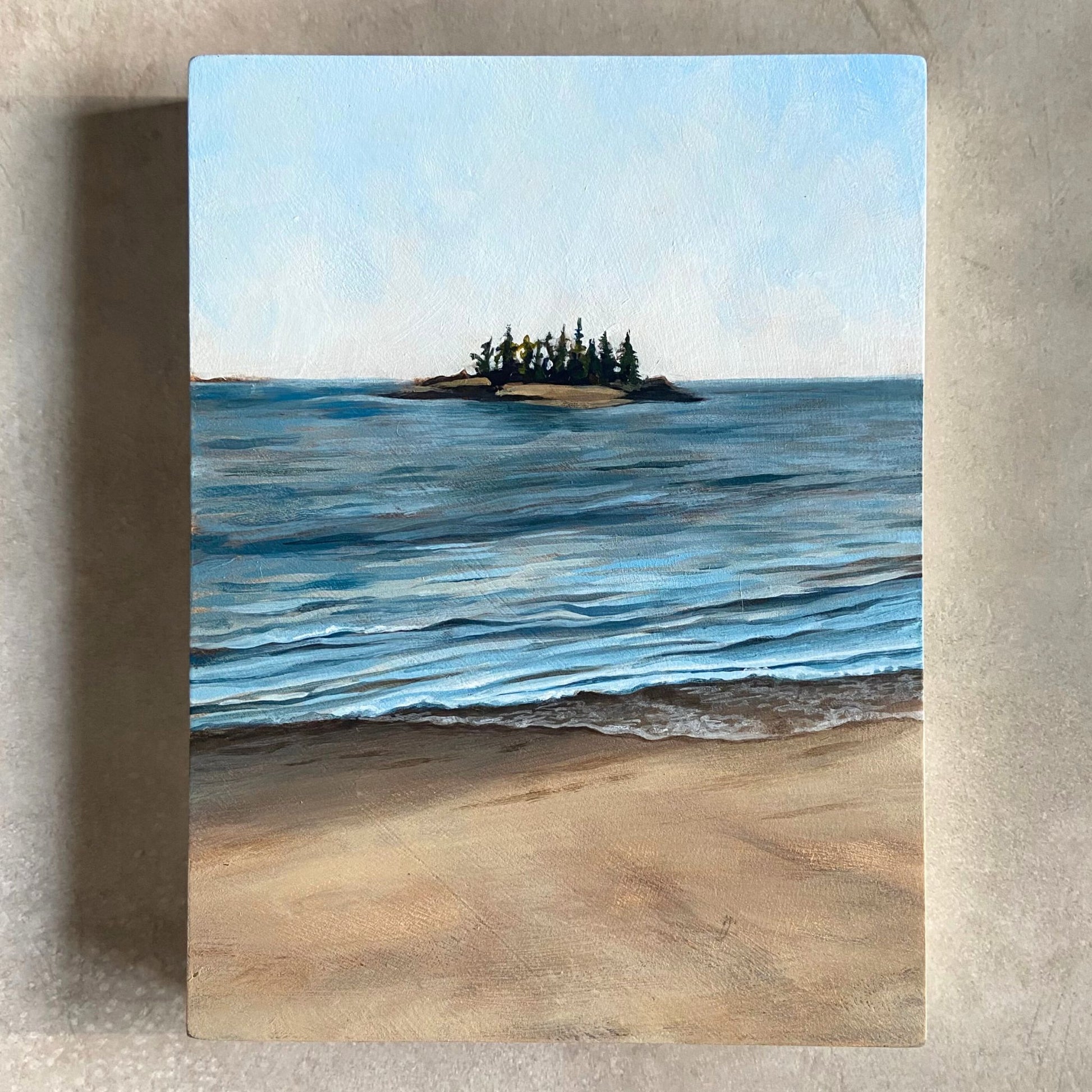 An acrylic painting on wood panel, sitting on a beige, textured background. The artwork, portrait-oriented, shows the sky at top, ocean waves in center, and a sandy beach at bottom. Centered on the horizon is a small forested island. 