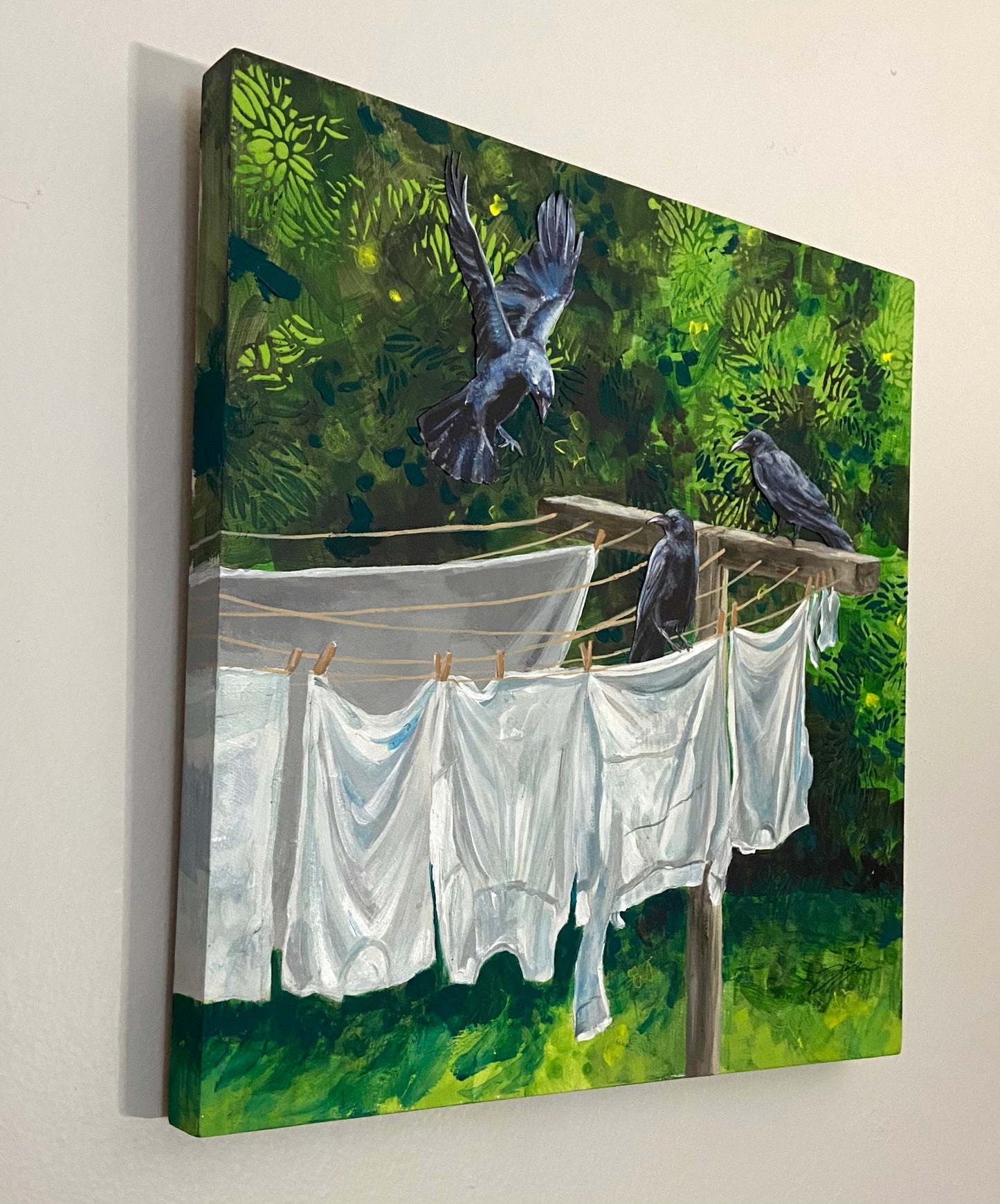 A view from an angle of a primarily green painting showing three crows at a clothesline of white laundry. The crows are collaged on paper.