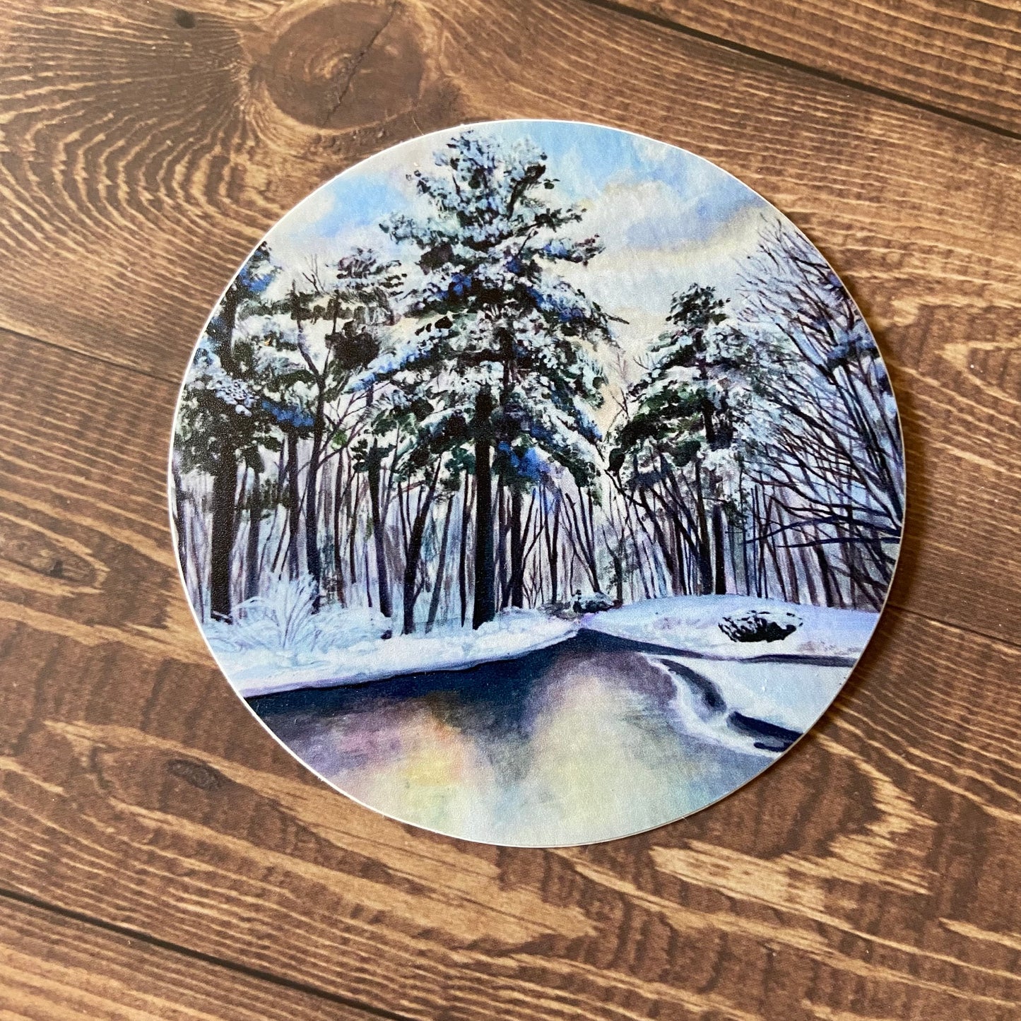 A round sticker of snow-covered trees by a road or water, sitting on a wooden surface. The decal is mostly light blue with other pastel colors and dark forest green in the pine trees. Watercolor style landscape painting sticker