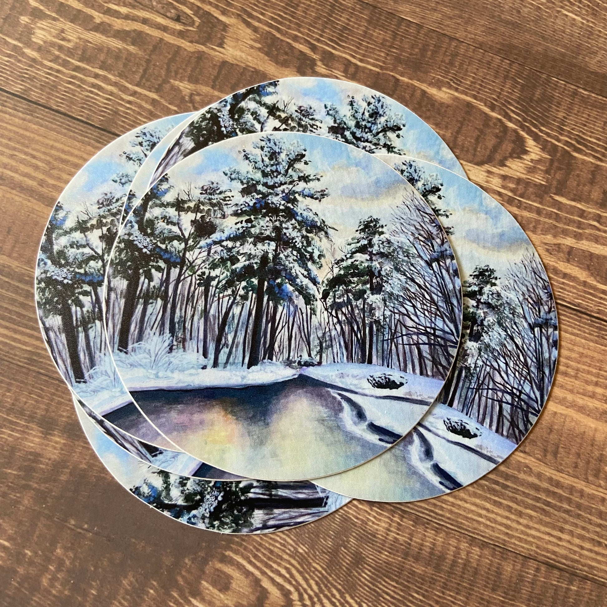 A small pile of round stickers showing a gouache painting of a winter landscape, sitting on a wooden surface. The art decal depicts trees, covered in fresh snow, around a river or wet road reflecting the morning light. Mostly pale blue and black.