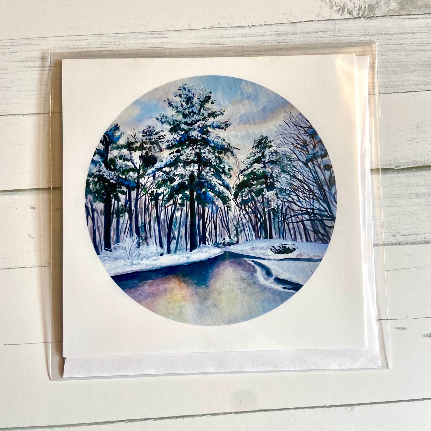 A folded white, square greeting card featuring my round painting, December 26, on the front. The painting is a predominantly blue snowy landscape, with snowy pines lining a still river. 