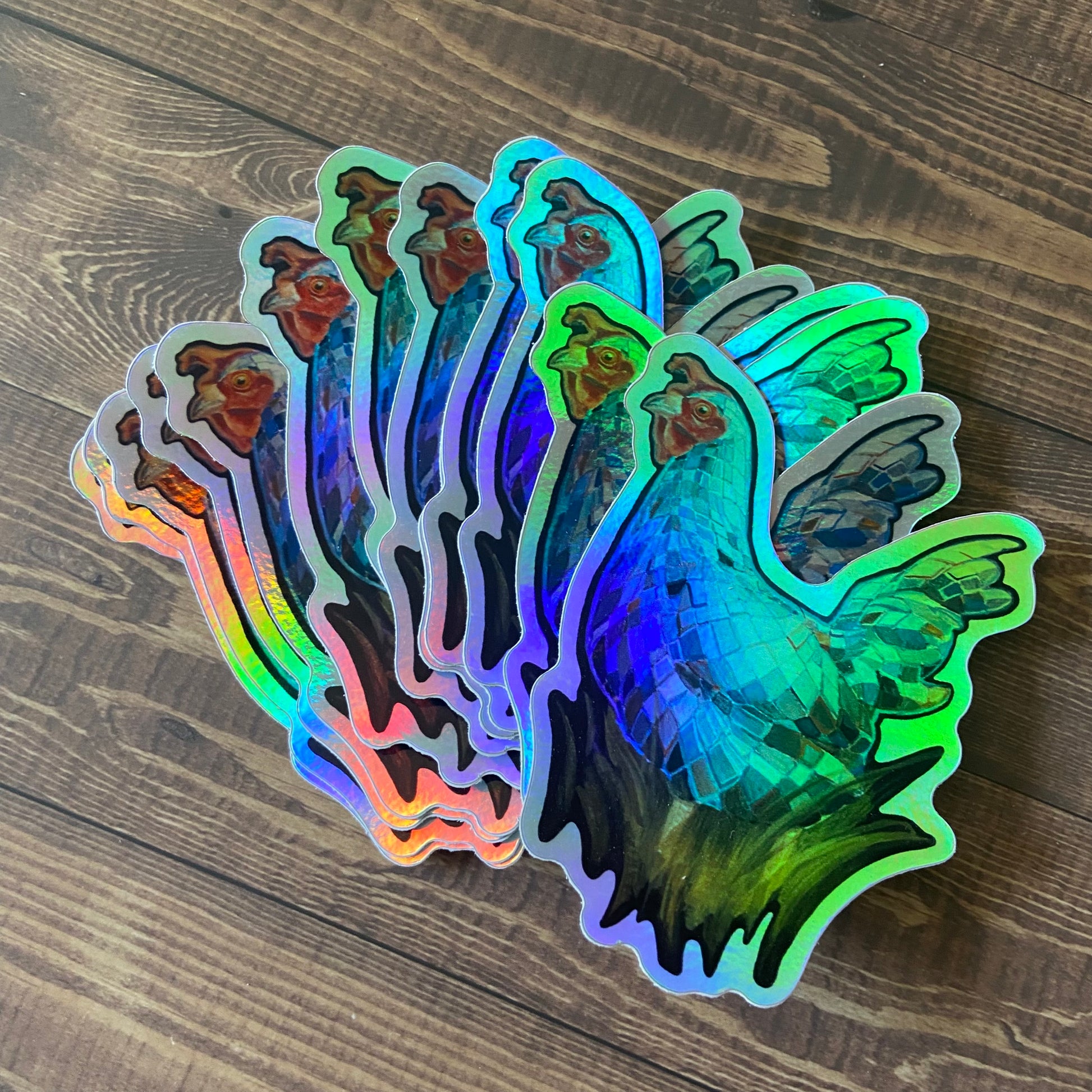 A pile of shiny stickers depicting a painting of a hen-shaped disco ball. The decals are printed on a glossy, holographic vinyl, making them look silvery yet reflecting a spectrum of colors. The stickers are sitting on a dark wooden surface.