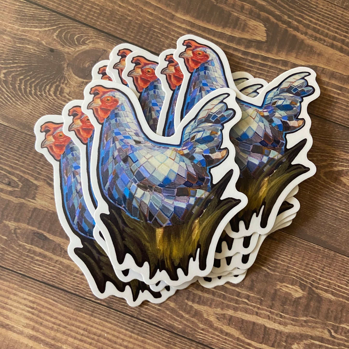 A stack of matte stickers depicting a chicken in profile, covered in mirror tiles like a disco ball. The hen stands in tall, dark grass. Each vinyl decal has a narrow white border and is die cut to the chicken's shape. They sit on a wood surface.