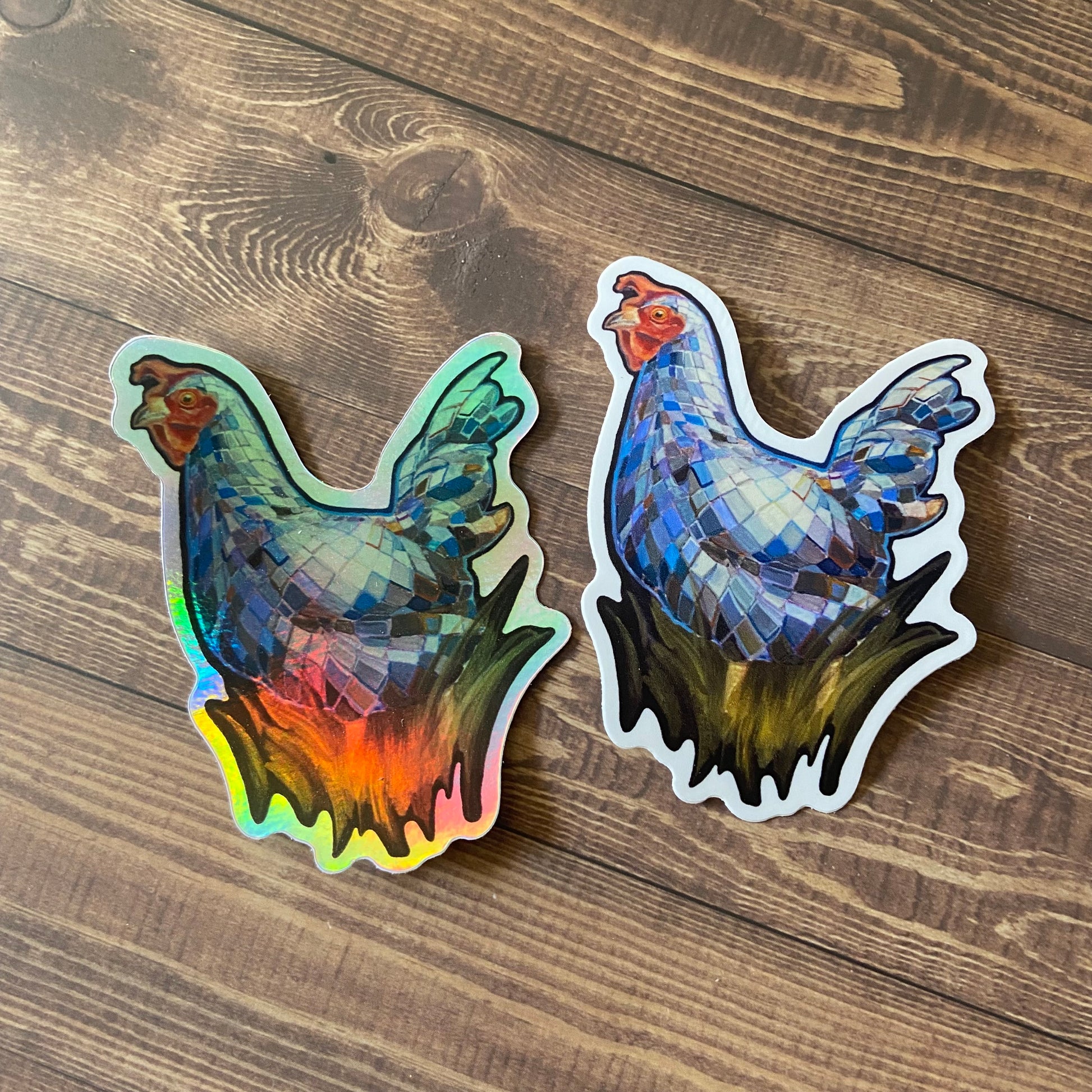 Two stickers of a painted chicken with a blue mirrorball pattern in tall dark grass. One sticker is holographic and glossy, reflecting rainbow colors. The other is standard matte with a thin white border. The stickers are on a dark wood surface.