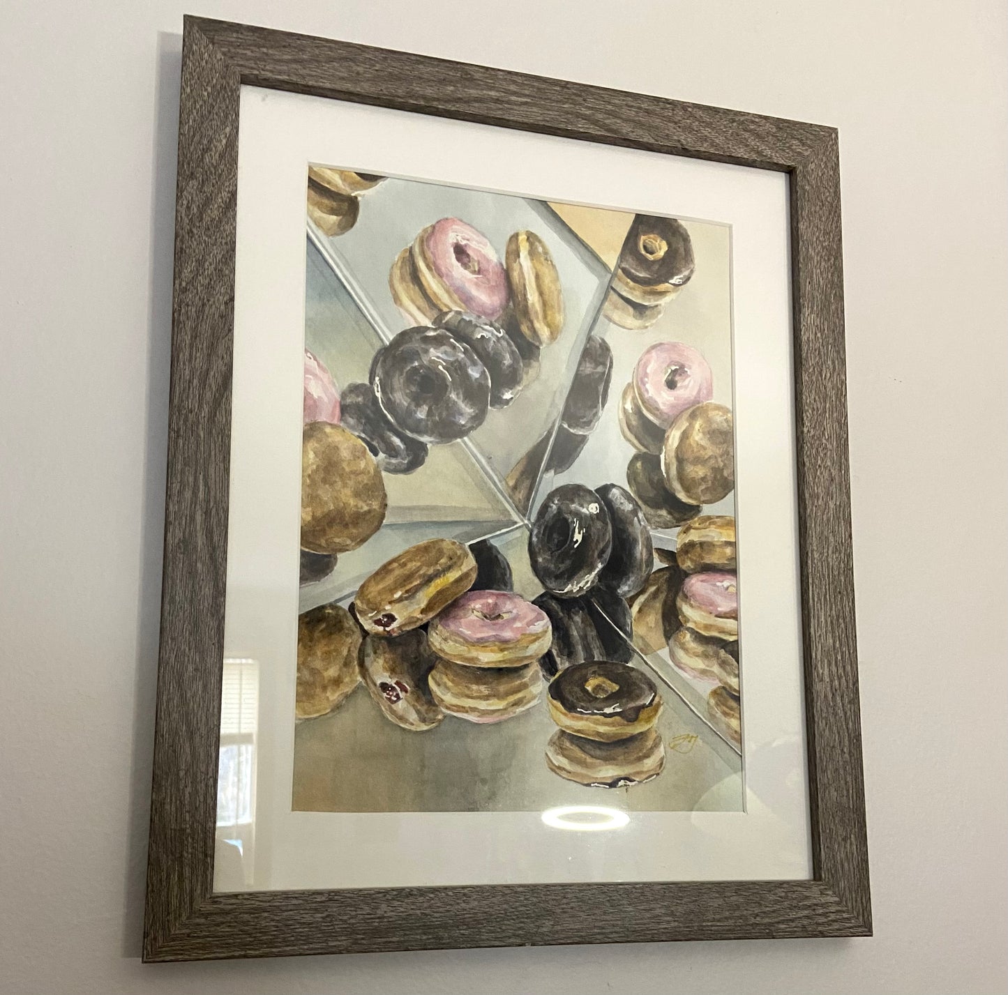 A watercolor still life painting of several varieties of donuts surrounded by mirrors, like a kaleidoscope. The painting is in a cool wood frame with a white mat. 