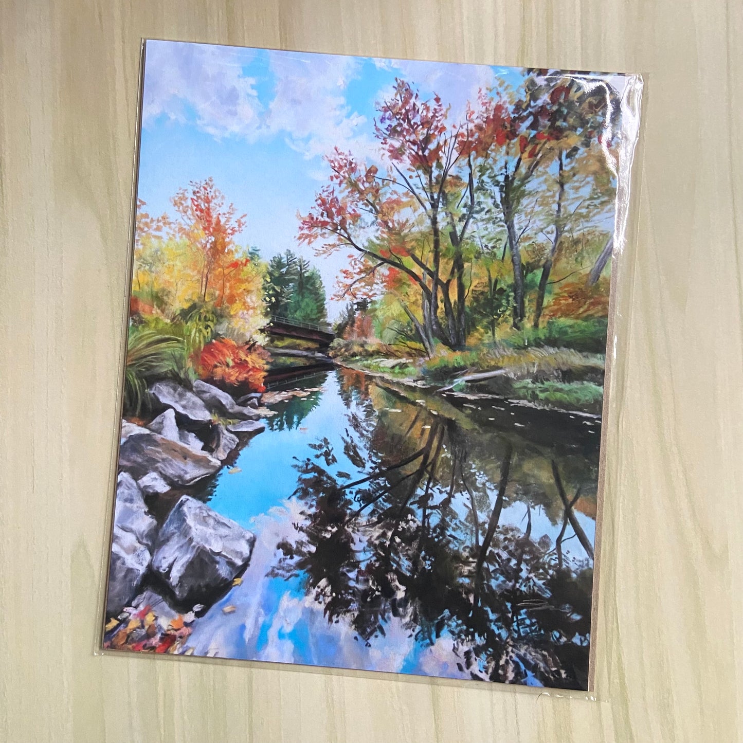 A river landscape art print showing autumn foliage reflected in the still water. The bright blue sky contrasts against the red, orange and yellow leaves. There is a bridge crossing the river at the far end of the scene.