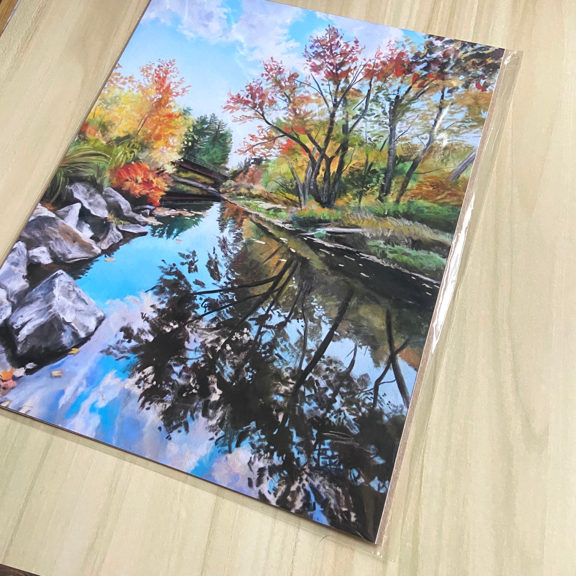 An angled detail view of the decorative art print. The chalky texture of the pastels can be seen, and the colors are vibrant and joyful. The fall leaves in the blue sky give a sense of serenity. Realistic gray rocks frame the edge of the water.