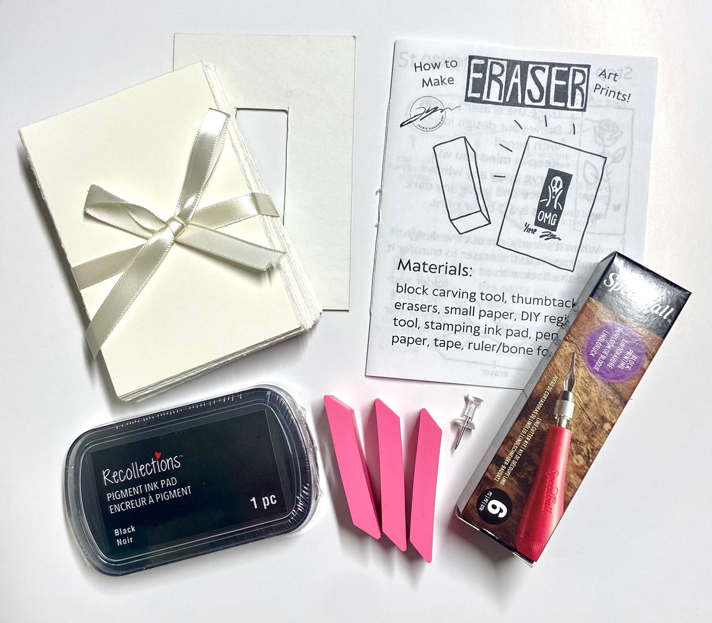 The contents of an eraser art printmaking kit laid outside of their package. Included are a registration tool, paper, an instructional zine, a black ink pad, three pink erasers, a thumbtack, and a linocut carving tool. 