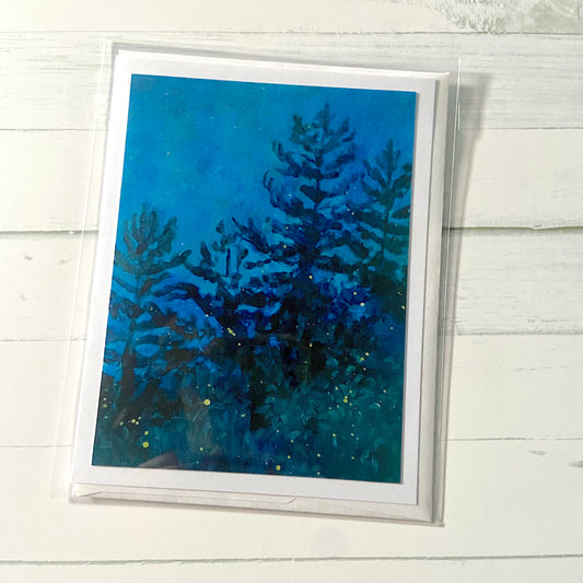 A folded white greeting card showing a nocturne painting of pine trees against a deep blue sky, fireflies' glow speckling the foreground.