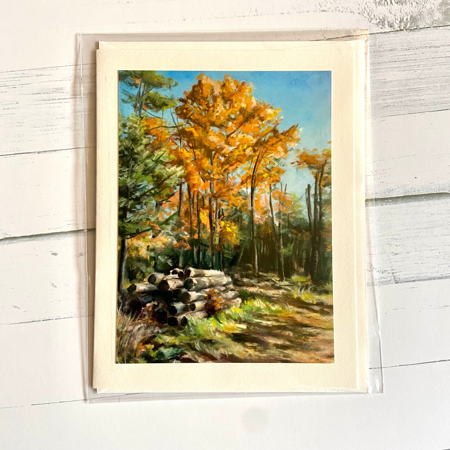 A folded cream greeting card depicting my pastel painting, "Free Dry Wood, Pick Up Only." Tall autumn orange trees rise into a clear blue sky, while on the ground in front of them is a pile of cut dry logs.