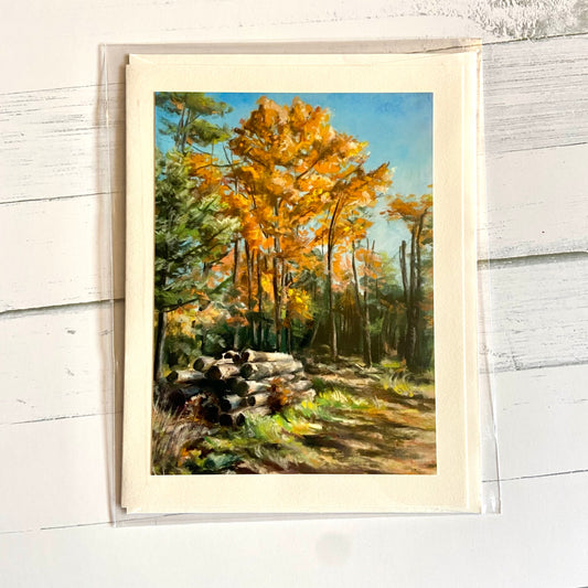 A folded cream greeting card depicting my pastel painting, "Free Dry Wood, Pick Up Only." Tall autumn orange trees rise into a clear blue sky, while on the ground in front of them is a pile of cut dry logs.