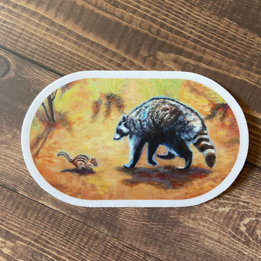 An oval-shaped orange sticker on a wooden surface. The decal shows a gouache painting of a raccoon and chipmunk forest encounter- the woodland creatures are making friends!