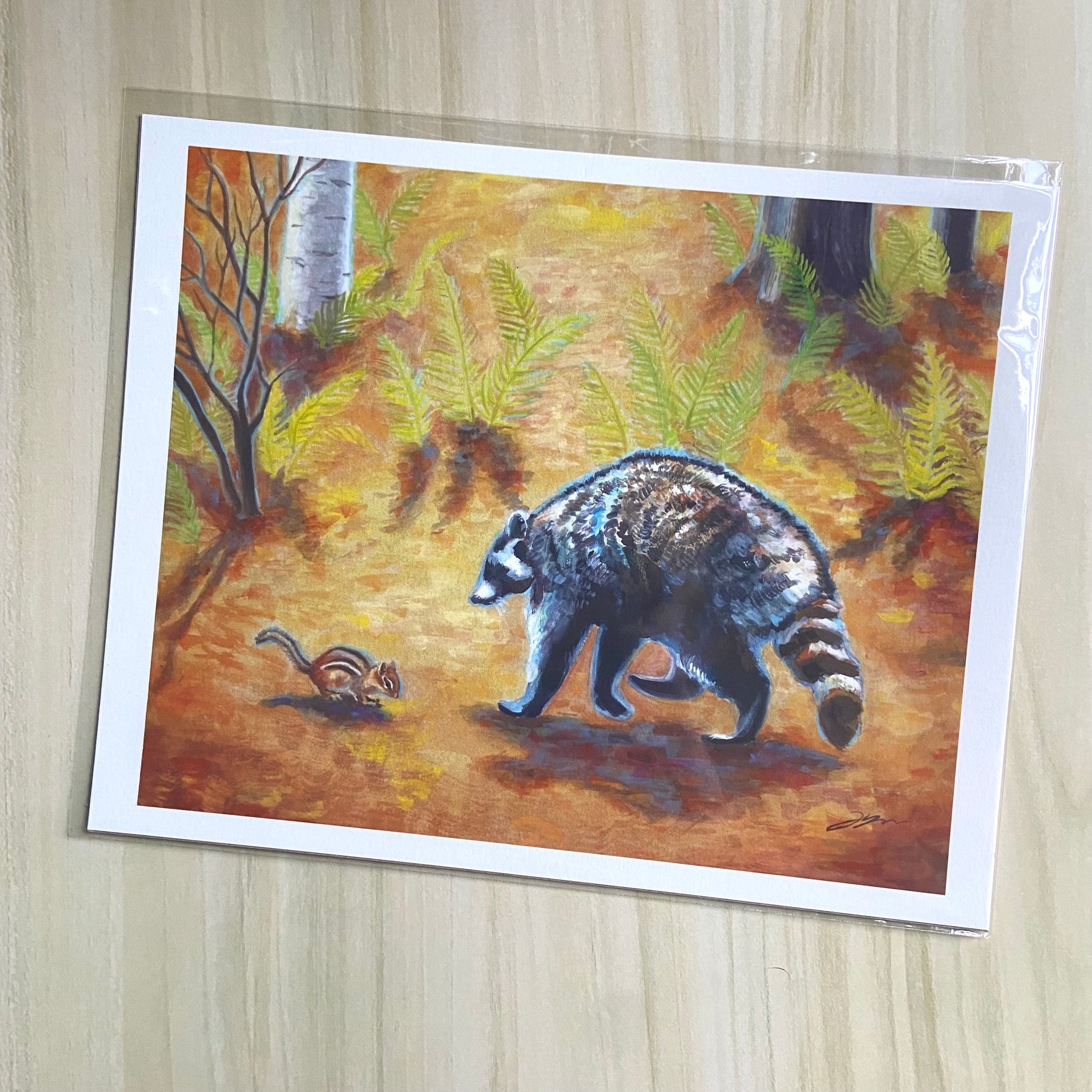 An orange art print of a raccoon and chipmunk meeting in the forest. The forest floor is covered in orange leaves and ferns. Gouache painting with visible texture. Print has a narrow white border and is wrapped in a protective sleeve.