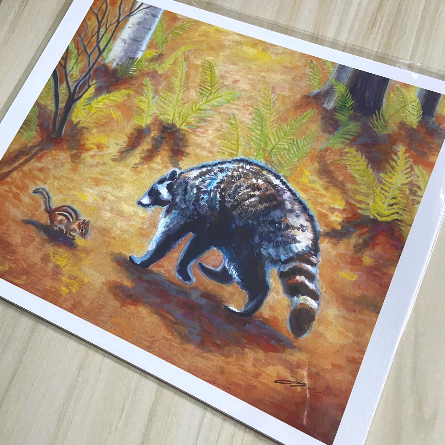 An angled detail view of the woodland creatures art print. The vibrancy and texture of the gouache painting is clear in the print, especially in the raccoon's fur. This fun fall artwork has a warm feeling.