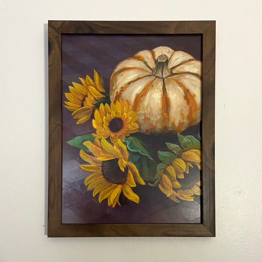 A still life painting of a striped pumpkin and several sunflowers on a purple plaid tablecloth. The painting is in a dark wood frame. 