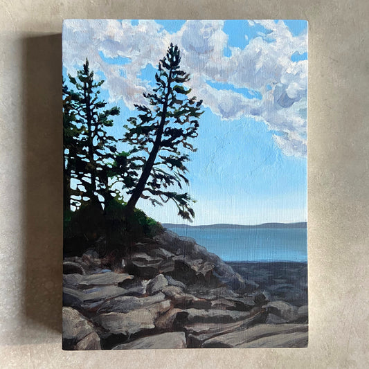 An acrylic landscape painting on wood panel, sitting on a beige, textured background. The artwork, portrait oriented, depicts a couple of pine trees growing above a rocky beach. A calm ocean bay is in the background and the sky is blue with white clouds. 