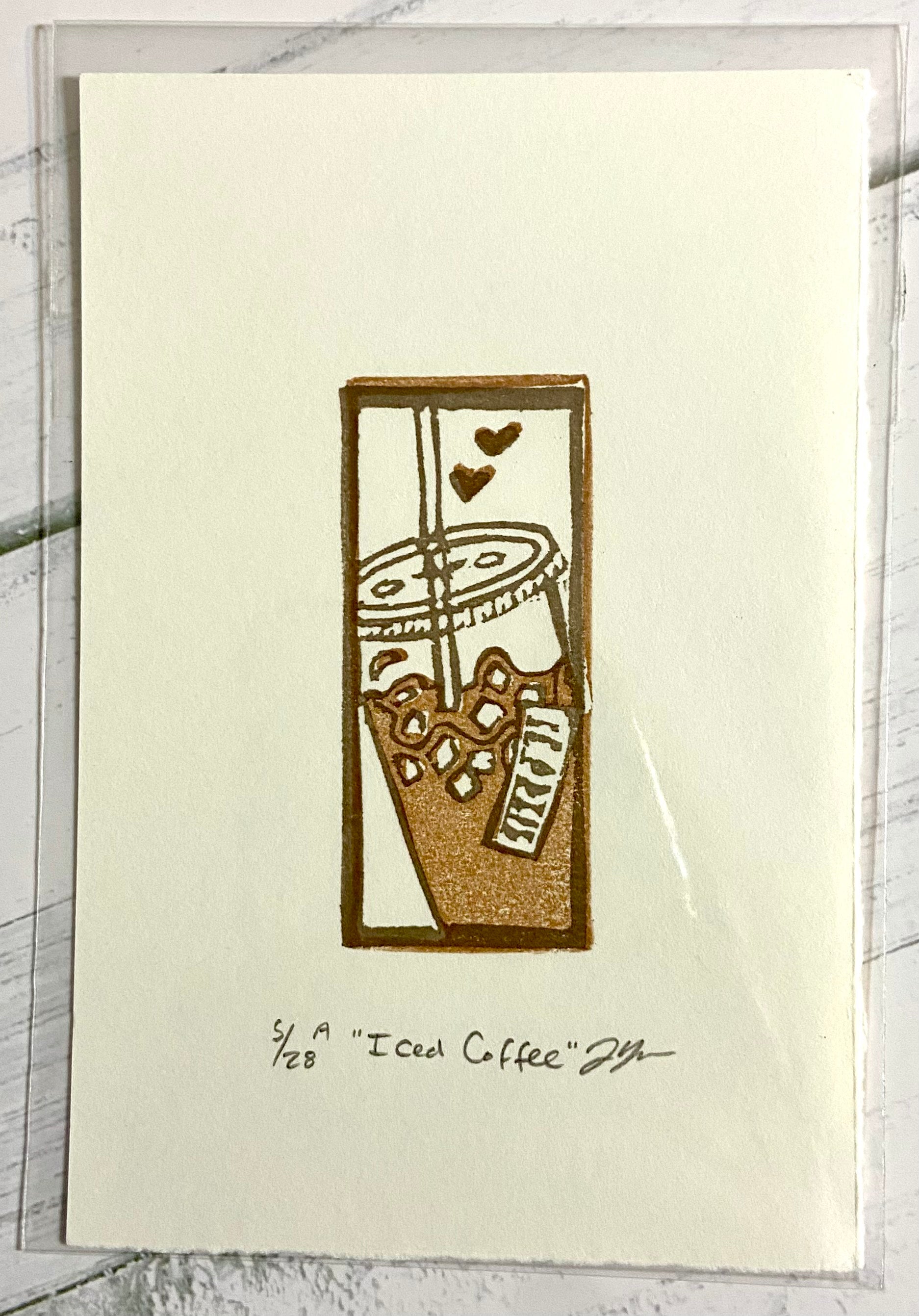 An image of a small two-layered relief print depicting an iced coffee in two shades of brown. the print is made by hand from a carved eraser and is numbered, titled and signed. 