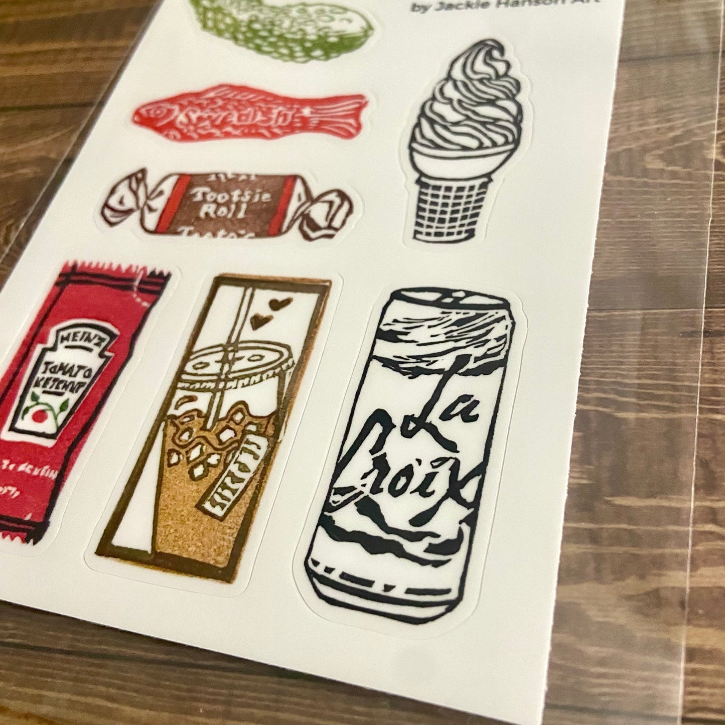 A close-up angled view of the  eraser art sticker sheet. Individual cut lines around the 7 stickers can be seen. The stickers are each around 1x2 inches and include a pickle, Swedish fish, tootsie roll, ice cream, ketchup, iced coffee, and La Croix.