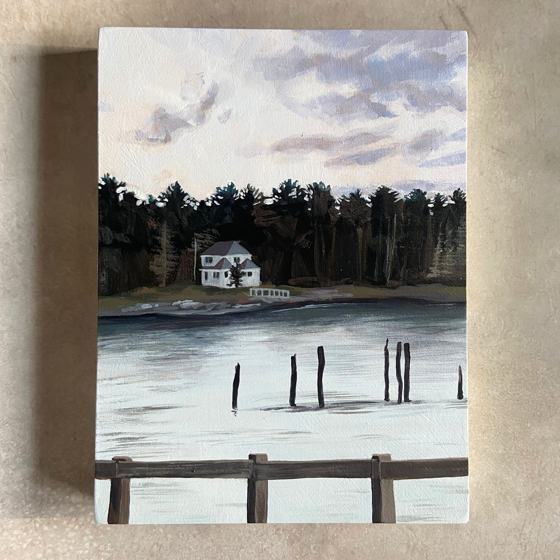 An acrylic landscape painting on wood panel, sitting on a beige, textured surface. The painting depicts a body of water reflecting an overcast sky, with a wooded piece of land separating the two. There is a gray house on the shore, remains of an old pier in the water, and a fence at the bottom of the composition. 
