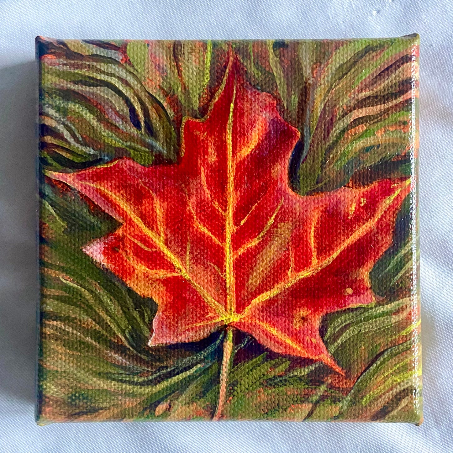 A square painting of a red leaf on green and brown grass. 