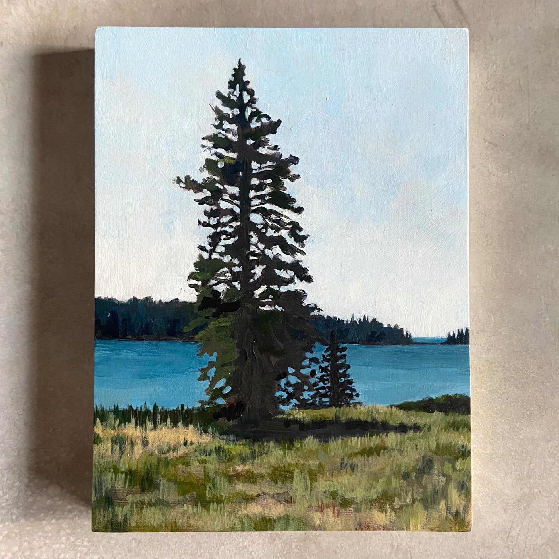 A landscape painting on wood panel, sitting on a beige, textured background. The artwork, portrait-oriented, focuses on a tall pine tree growing on a grassy field by an ocean bay. 