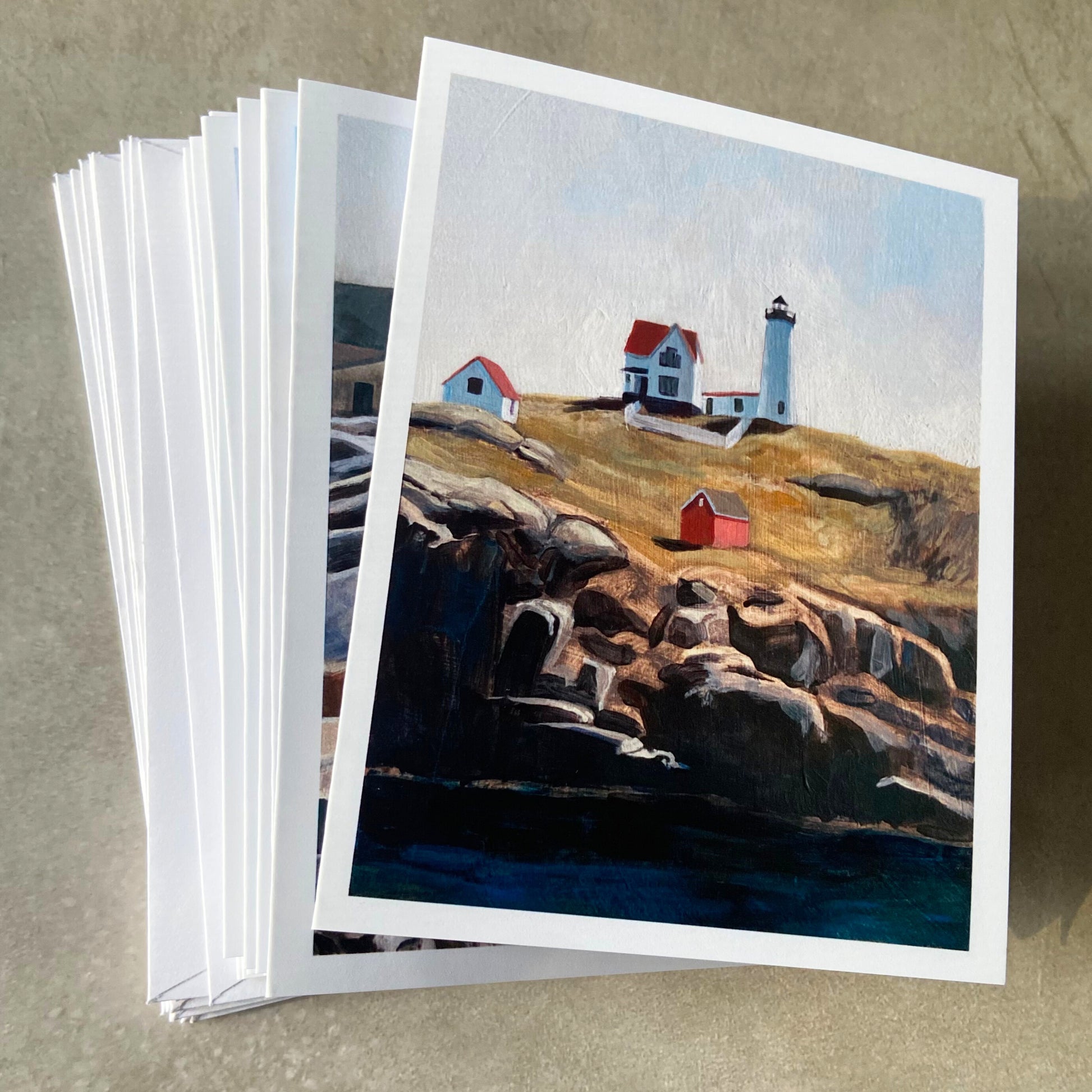 A stack of folded white greeting cards and envelopes on a gray surface. The card on top shows a lighthouse on a hill over a rocky cliff and deep blue water. Part of the next card shows at left and it is primarily blue/grey.