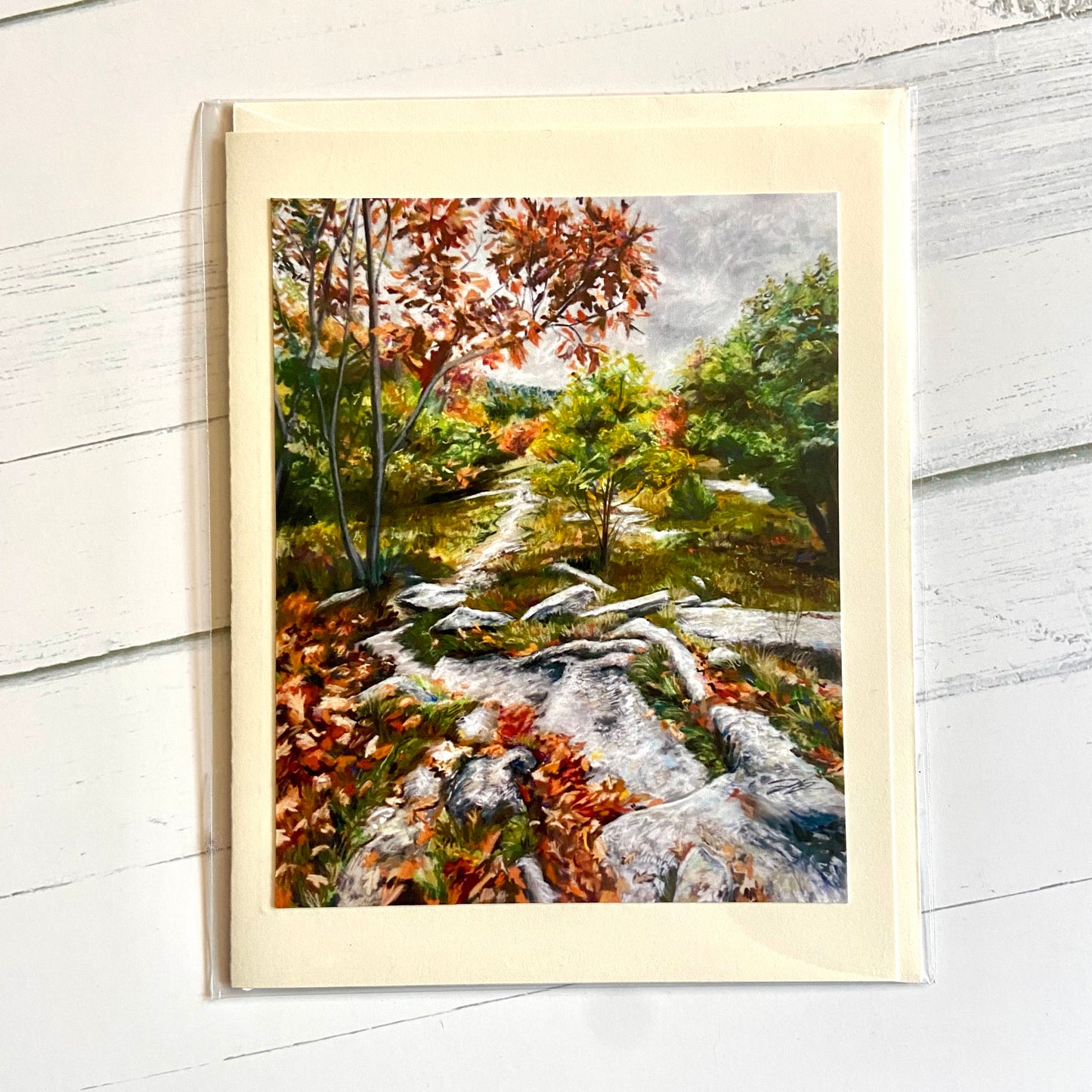 A folded cream-colored greeting card with an image of my pastel landscape painting, Looking Back: Mt. Rowe. A rocky path winds between late autumn leaves on the ground and in the trees, underneath an overcast sky. 