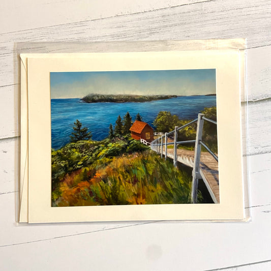 A landscape-style greeting card showing a painting of coastal Maine, looking down a lush hill at a wooden staircase, a red shed, and the Atlantic ocean. 