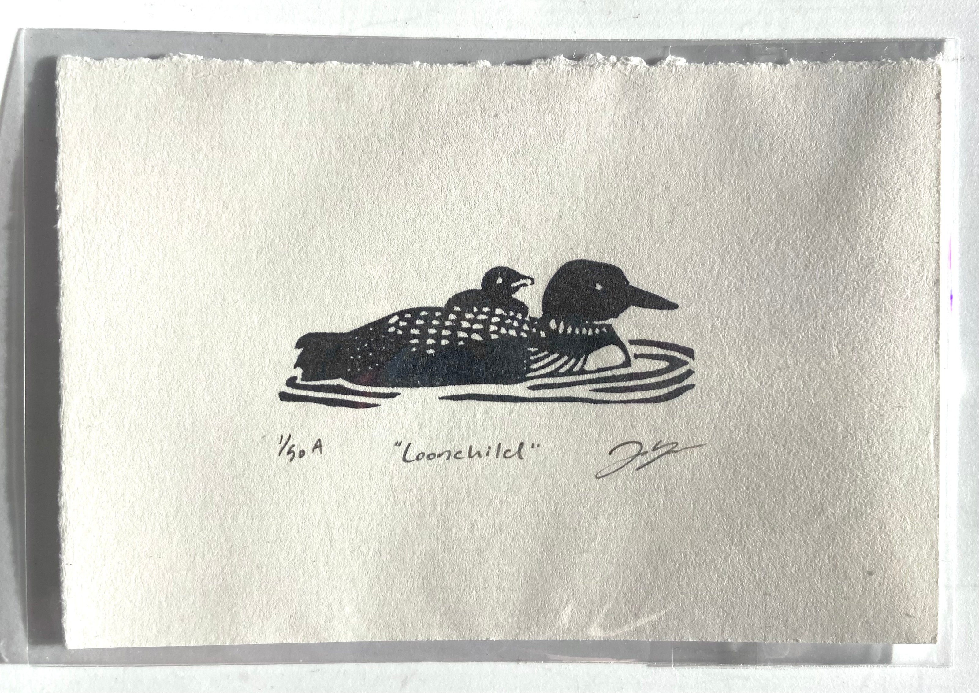 A tiny stamped print showing a loon with a baby on its back floating in water, with a wide paper border around it. Eraser printed are made by carving pink erasers and stamping them. The print is titled "Loonchild" and is signed and editioned. 