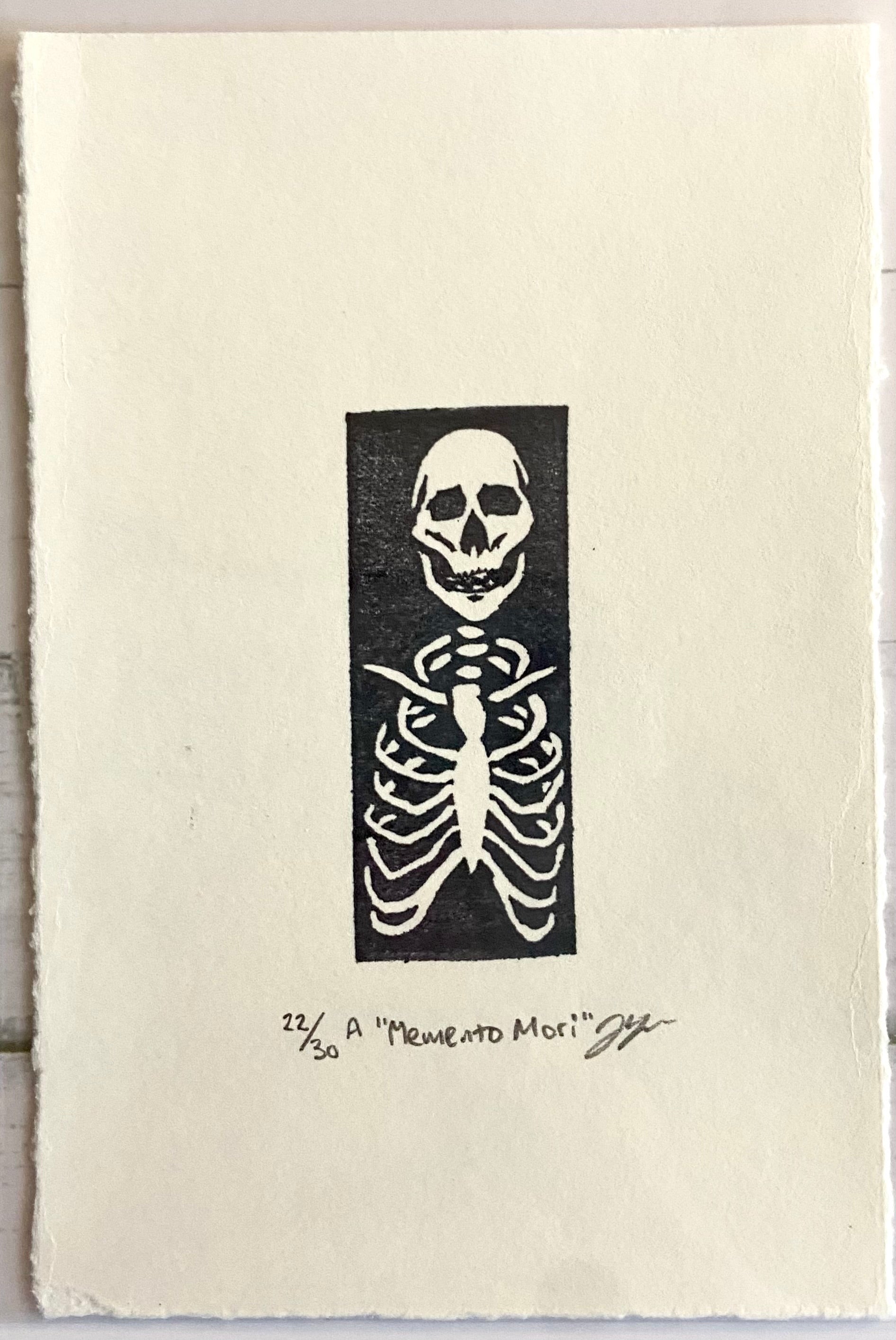 A tiny stamped print showing a skeleton's ribcage and skull in white on a black rectangle, with a wide paper border around it. Eraser printed are made by carving pink erasers and stamping them. The print is titled "Memento Mori" and is signed and editioned.