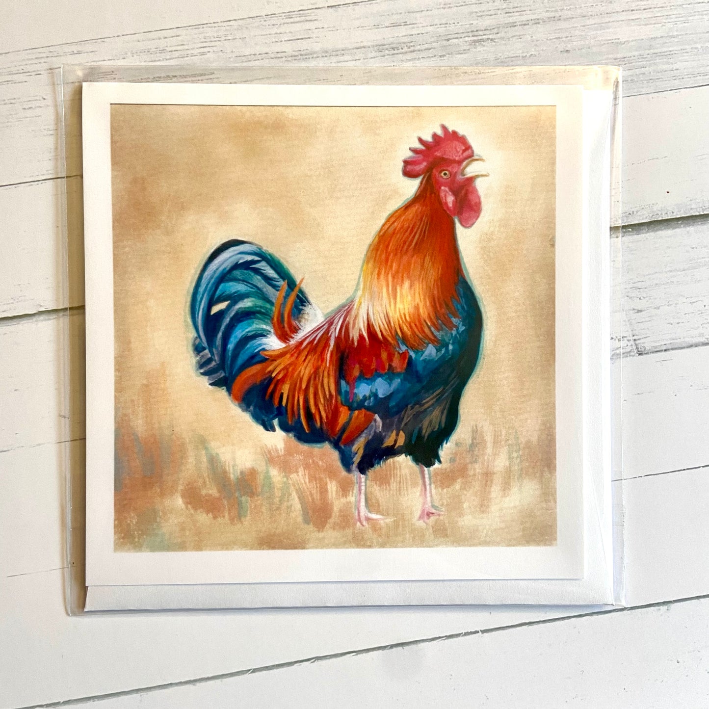 A square, folded white greeting card depicting my gouache painting of a rooster mid-caw. An orange and blue rooster stands in the center of the image, calling out with a blank stare in its eye. The background is beige and abstract.