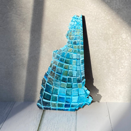A New Hampshire- shaped painting on wood, painted with the pattern of a blue disco ball. The painting is stood up in a patch of sunlight.